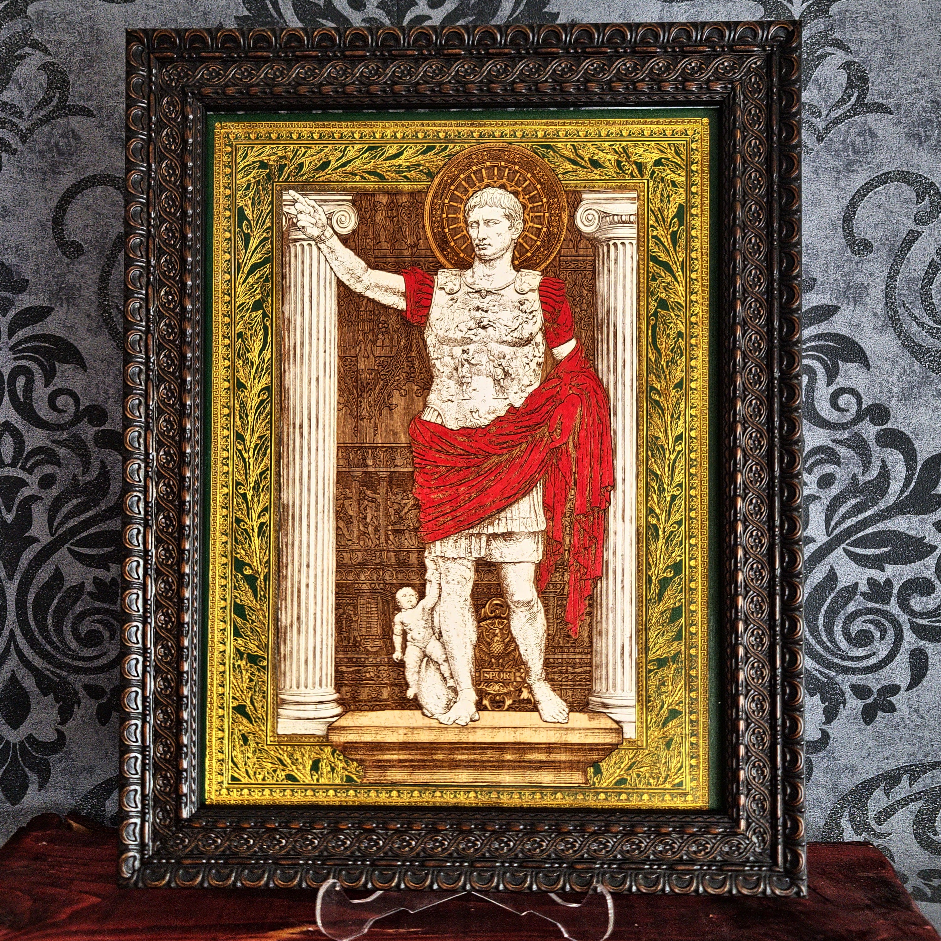 A framed art print depicting a Roman emperor standing before columns, with a red draped robe and a gold halo, set in an ornate dark frame against a patterned gray wall.