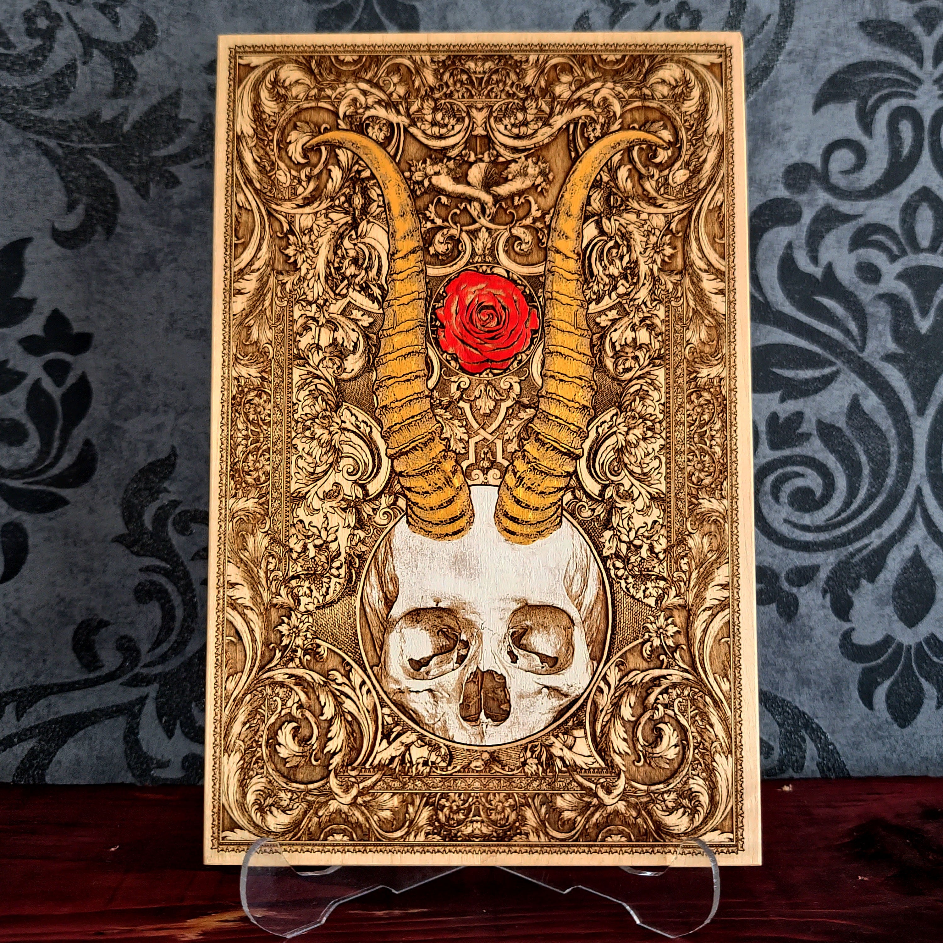 A rectangular decorative plaque or art print with an ornate gold-toned baroque-style design, featuring a skull at the bottom, large curved horns rising upward, and a red rose near the top center.