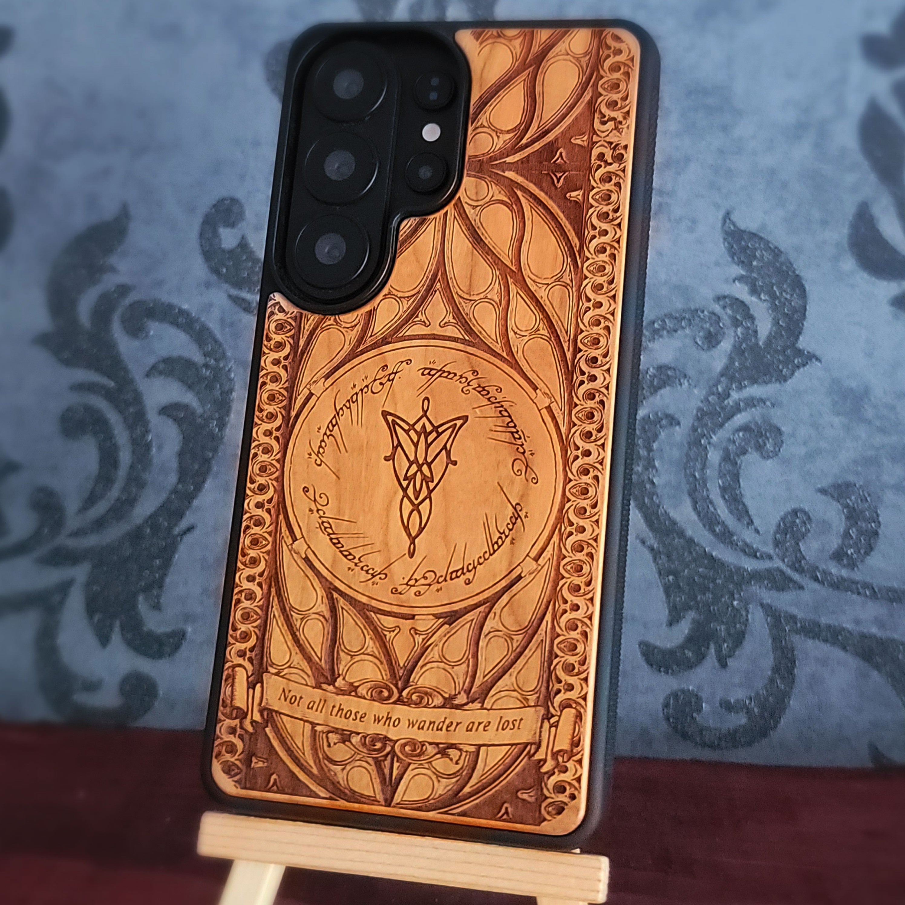A custom carved wooden phone case with intricate designs inspired by Lord of the Rings, shown standing on a small wooden display stand against a blue damask patterned background.