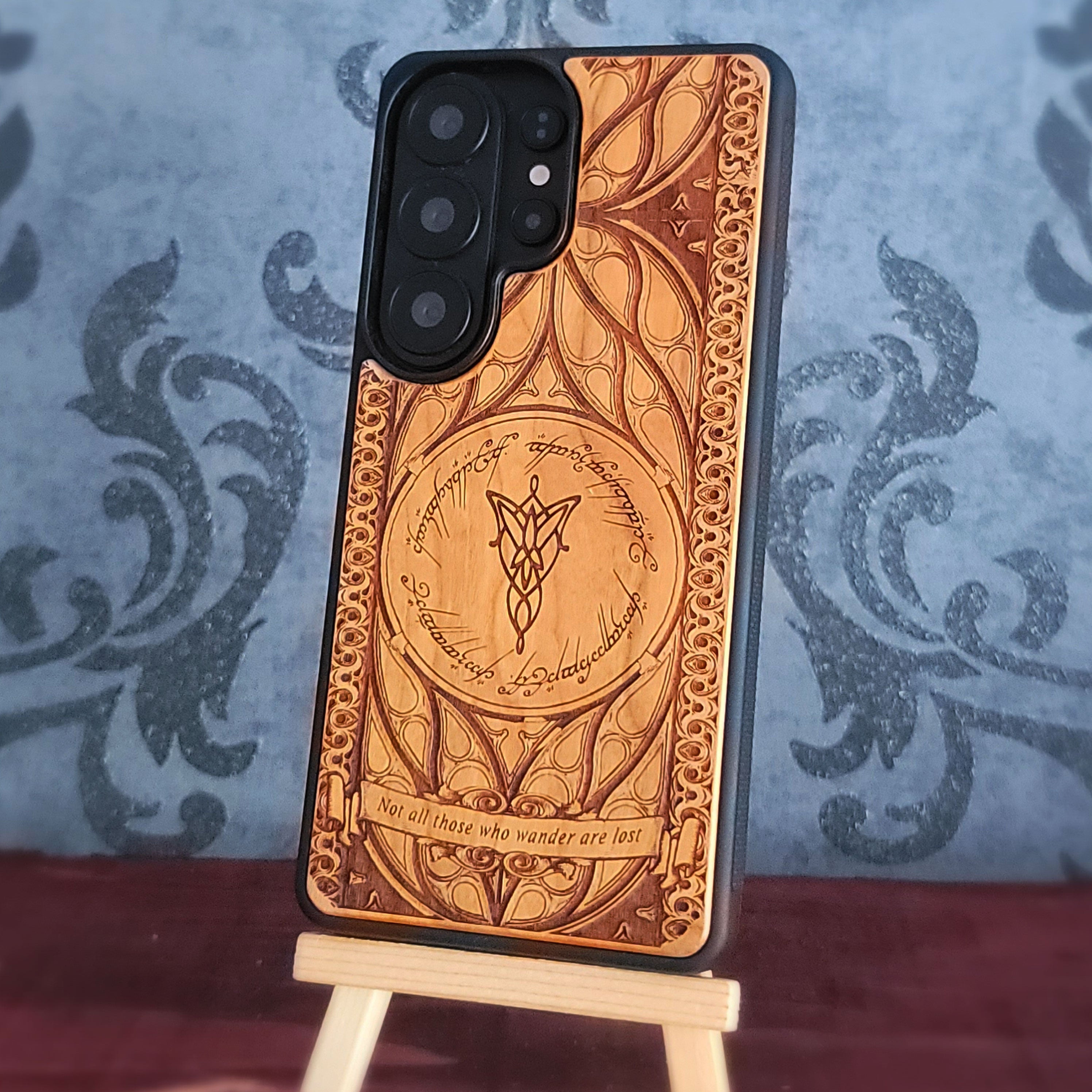 A phone case with an intricate carved wood design featuring a central ring motif and Elvish script, displayed on a small wooden stand against a blue damask patterned background.