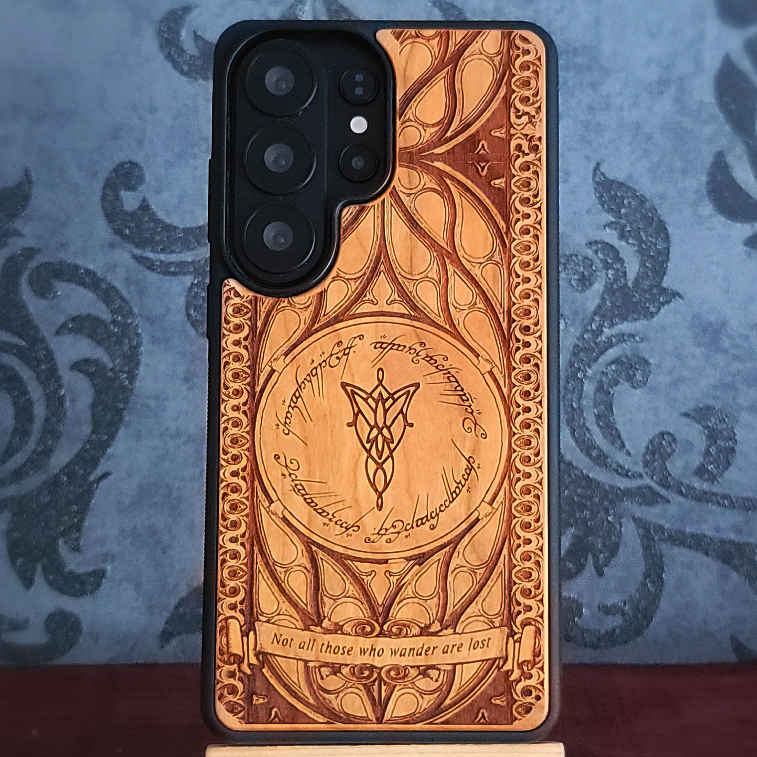 A wooden phone case with an engraved Hobbit-style door design, featuring Elvish script and a central symbol, shown on a smartphone. The case has a warm brown wood finish with darker burned details, set against a blue patterned backdrop.