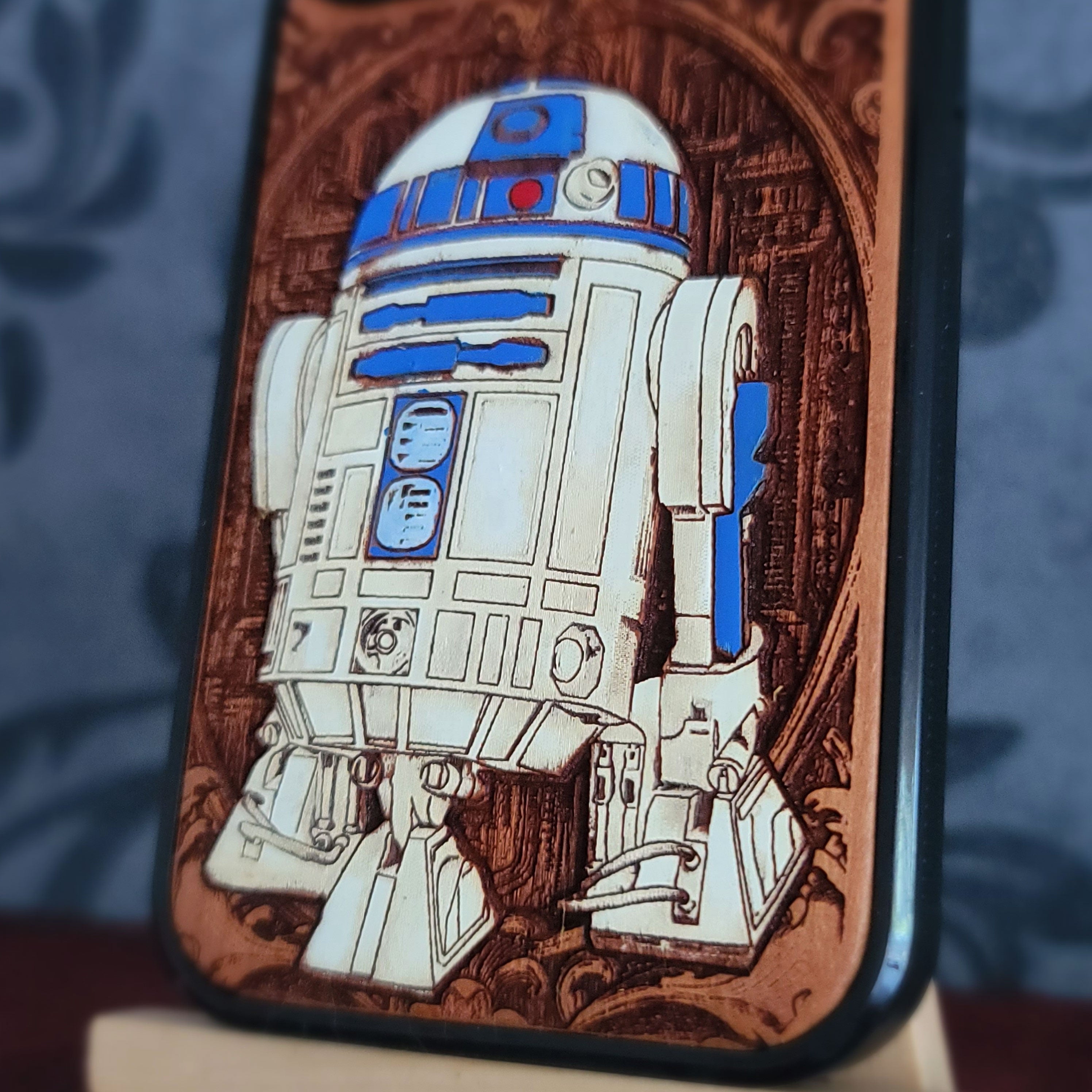 A close-up of a phone case featuring a detailed illustration of R2-D2, set against a dark blue patterned background.