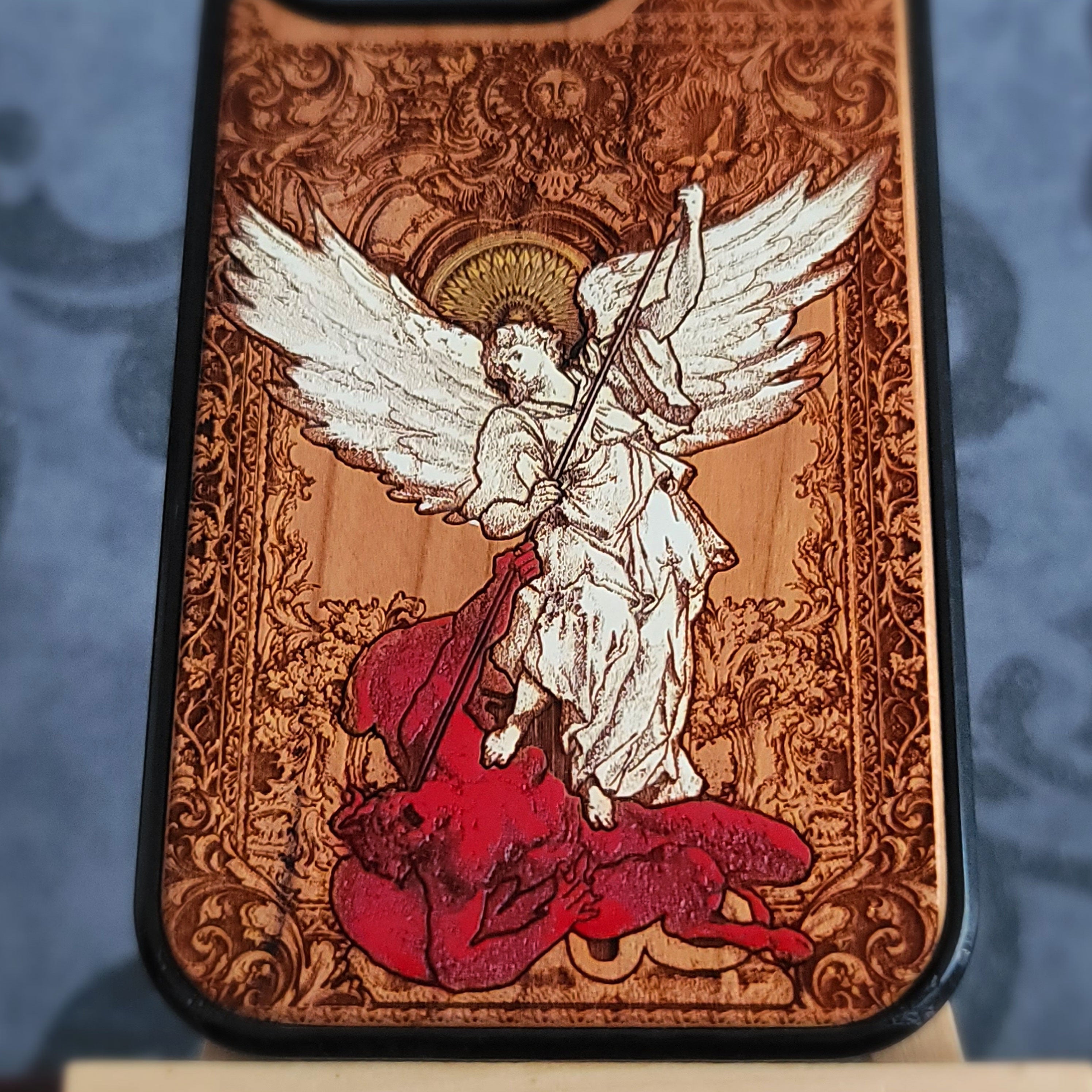 A close-up of a phone case featuring a detailed religious illustration of Saint Michael angel with large white wings standing on a red creature, set against an ornate brown baroque-style background.