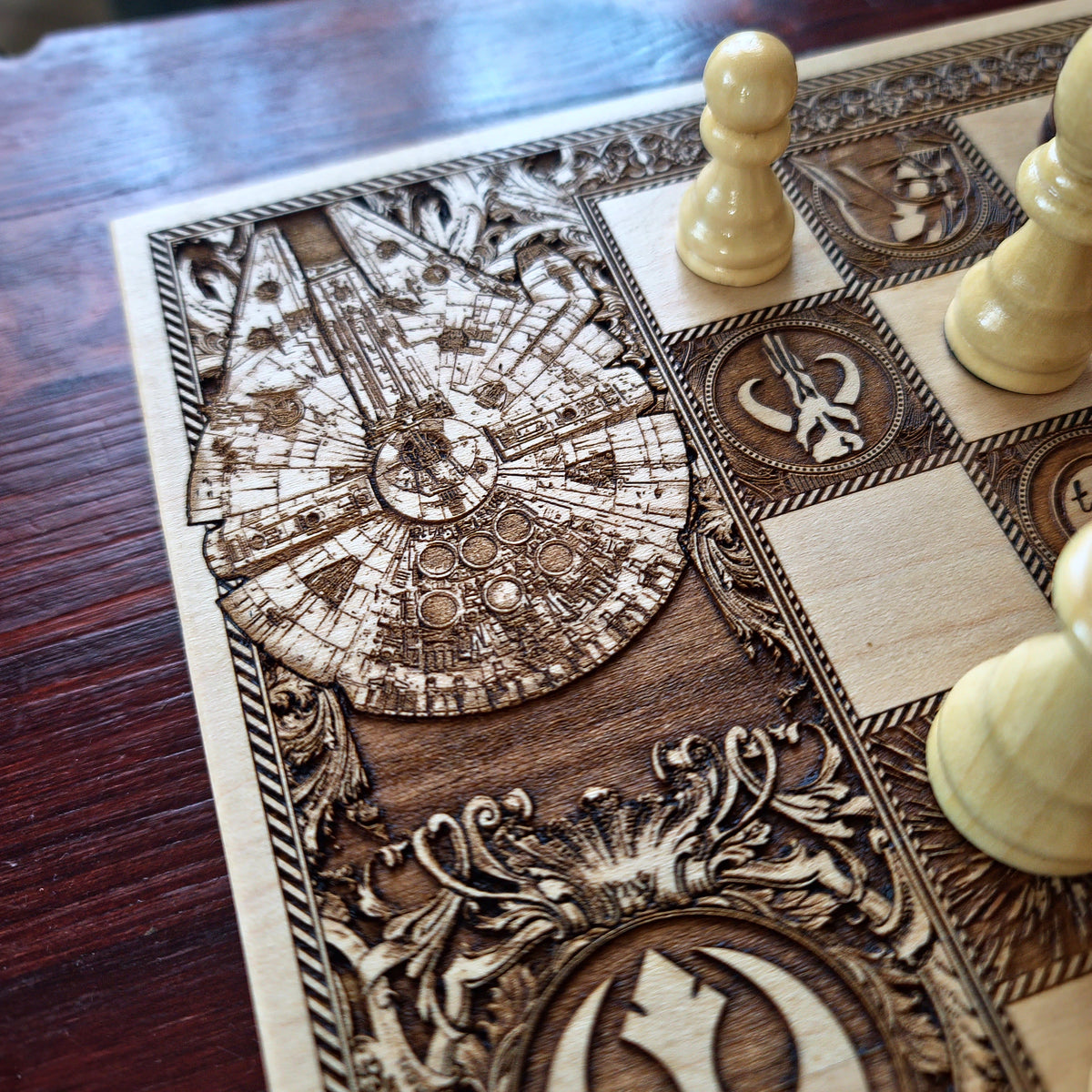 Close-up of an intricately engraved wooden chessboard featuring Star Wars-themed designs, including a Millennium Falcon and Rebel Alliance emblems, with light-colored wooden chess pieces on a dark wood tabletop.