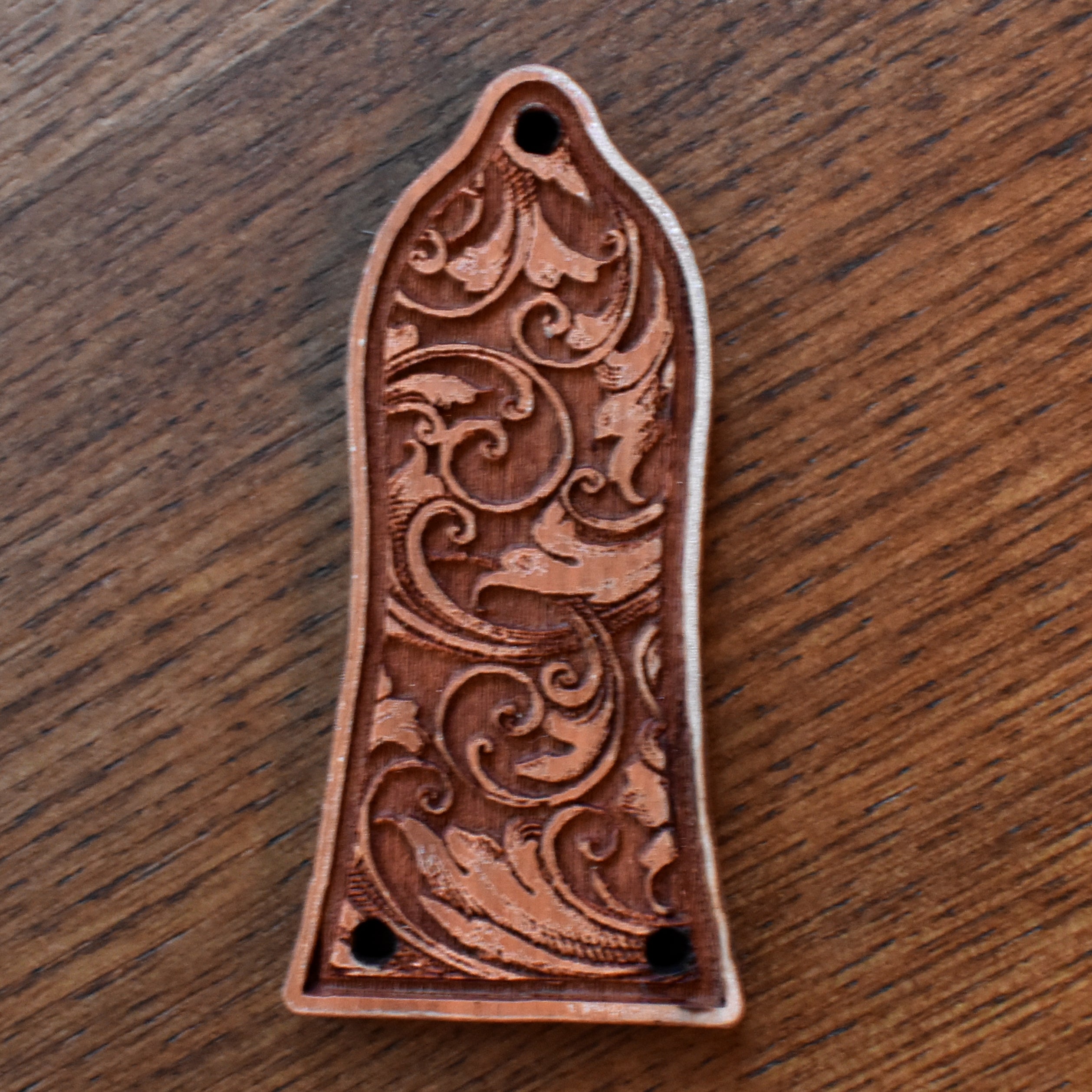Laserβengraved wood trussβrod cover for an Epiphoneβstyle guitar. The wood has a medium brown finish with subtle grain texture. The central engraving features an elegant band of floral scrollwork. Two mounting holes are visible at the bottom edge.
