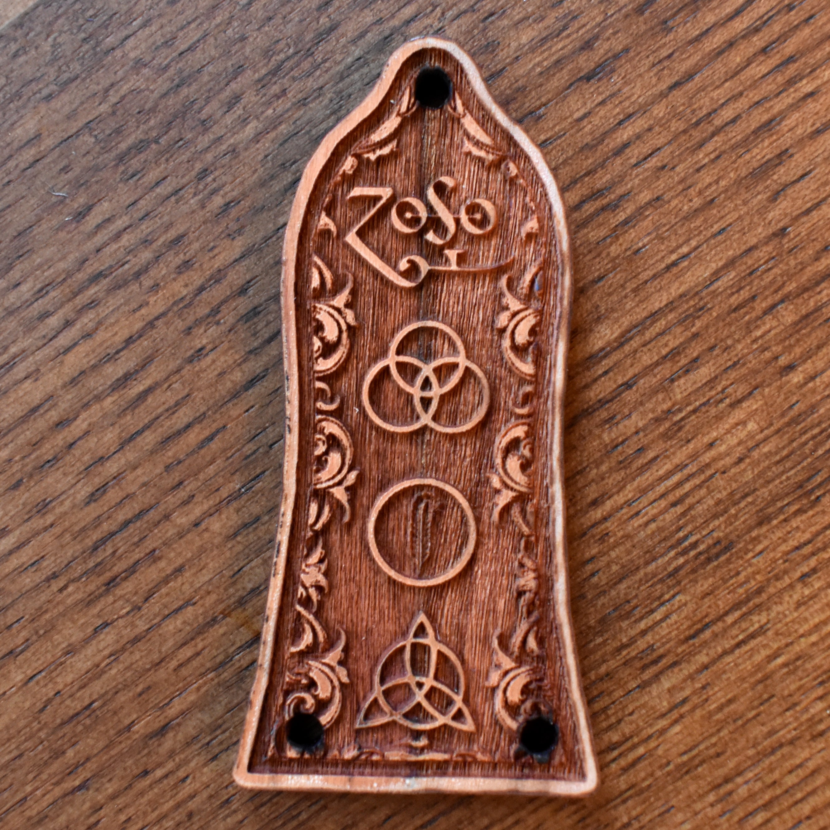 A laserβengraved wood trussβrod cover for an Epiphoneβstyle guitar. The wood has a warm reddishβbrown finish, and the cover is carved with ornate scrollwork flourishes along the sides. The centre features three Celticβstyle knot symbols: a trefoil knot at the top, a plain circle in the middle, and a triquetra at the bottom. Two mounting holes are visible at the bottom edge.