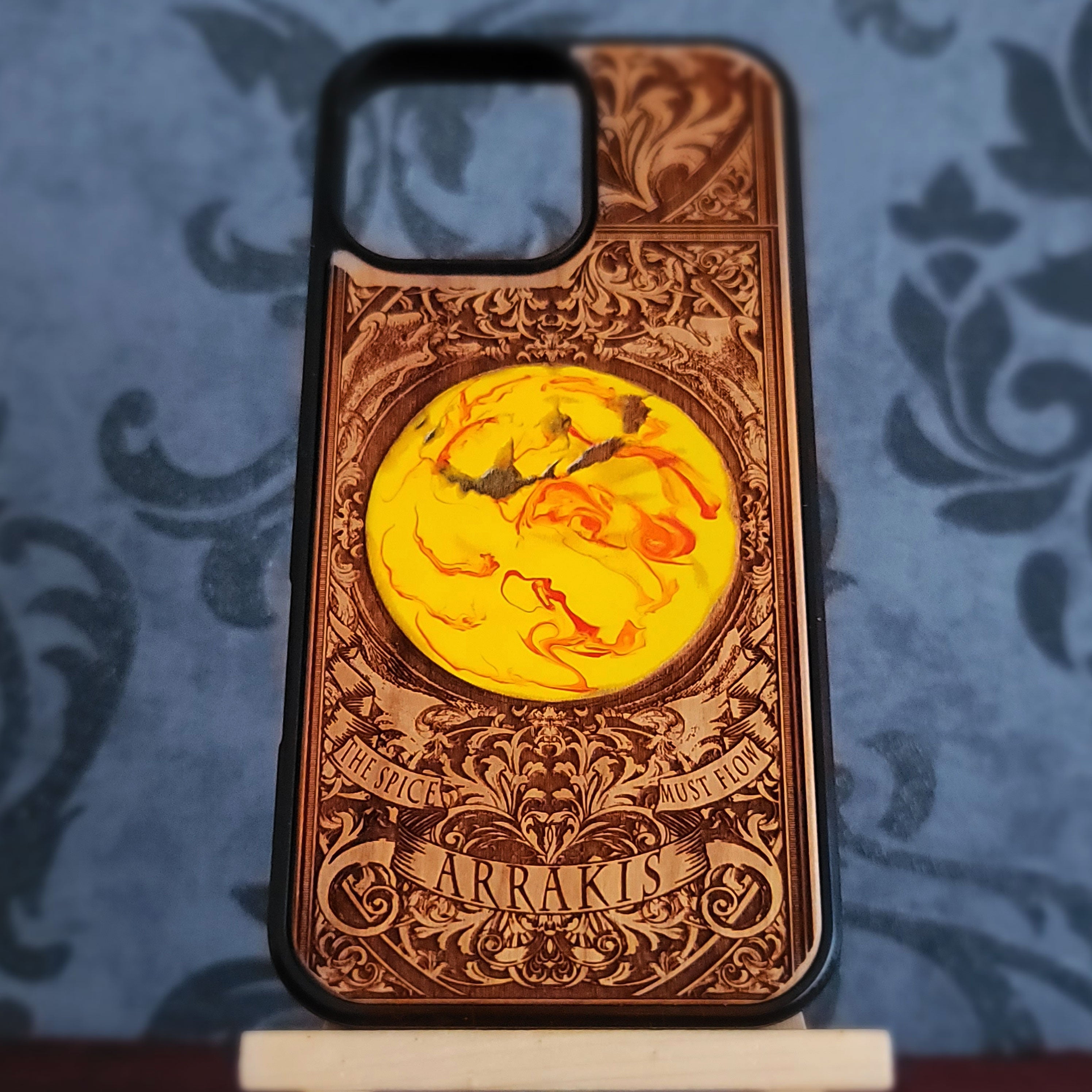 A custom wooden phone case with intricate laser-engraved baroque-style patterns, featuring a central circular yellow and orange marbled design and the name 'ARRAKIS' engraved at the bottom,