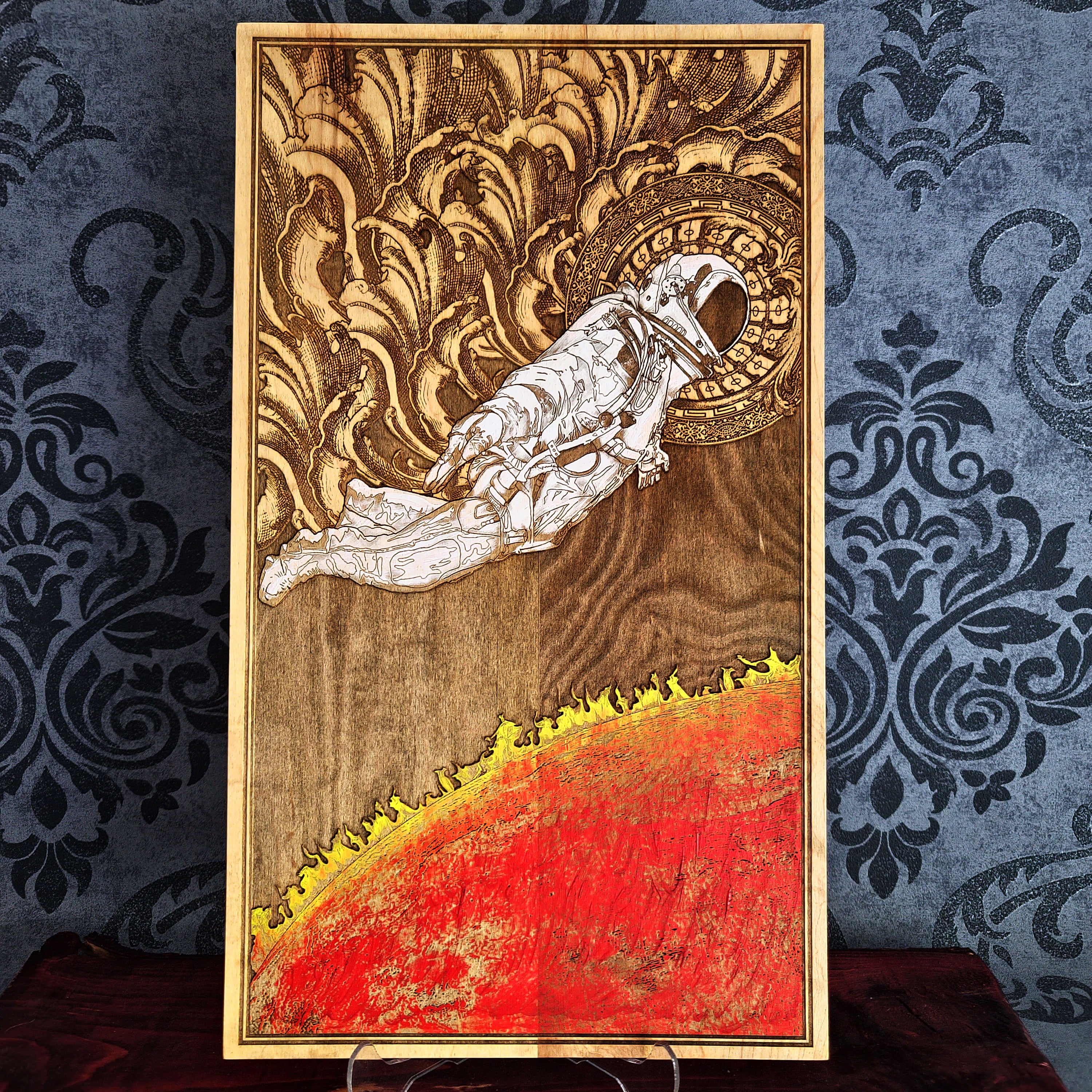 A wood burned artwork depicting an astronaut floating in space above a fiery red planet. The astronaut is wearing a white spacesuit and helmet. The background is a dark brown wood with intricate details.