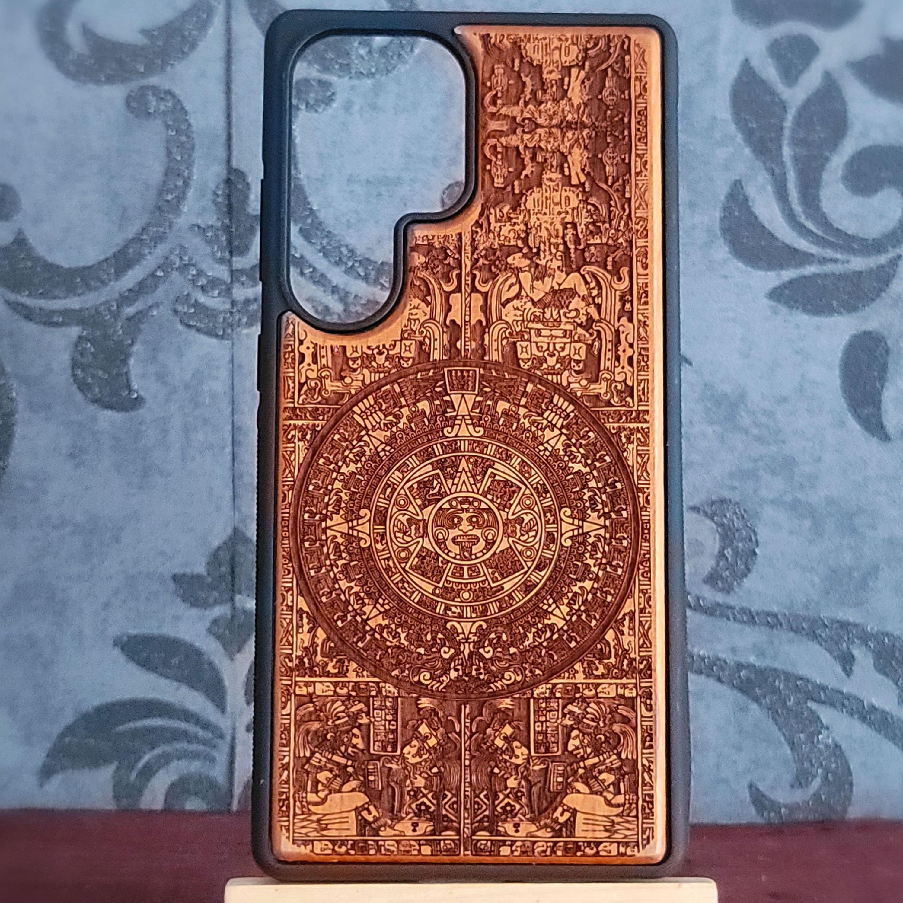 A smartphone case with a black bumper edge and a back panel featuring an intricate brown etched Aztec calendar design, displayed on a small stand against a blue patterned background.