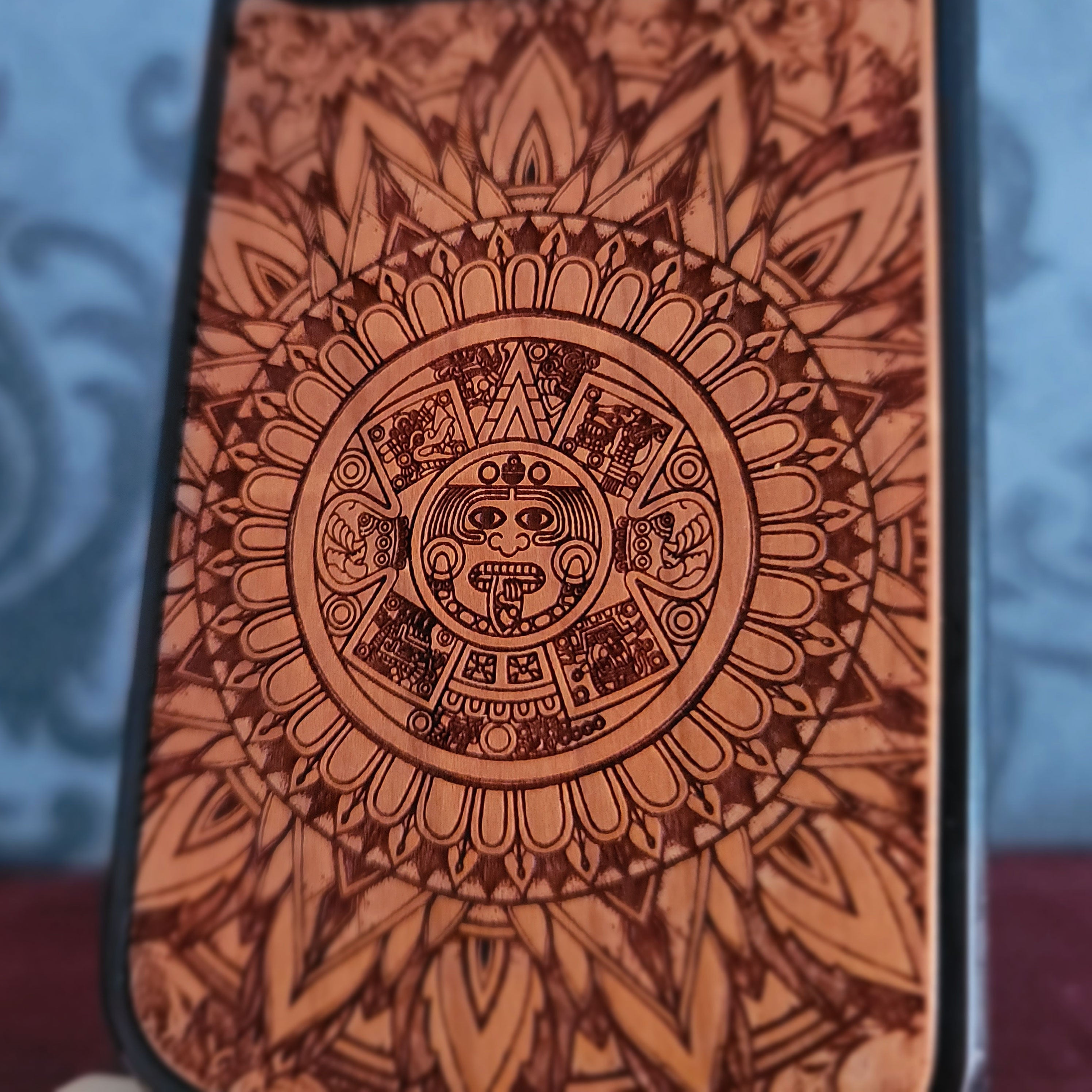 A close-up of a phone case featuring a detailed, circular Aztec calendar-style sun design in dark brown engravings over a warm orange-brown wood-like finish.