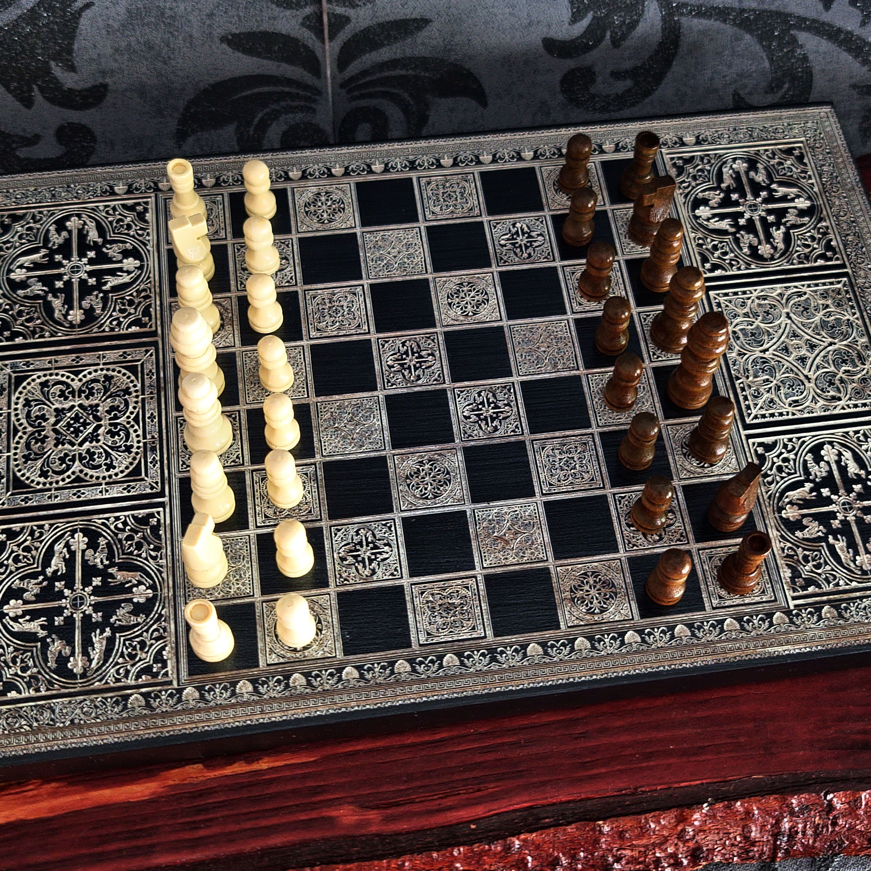 An ornate chess set displayed on a decorative chessboard. The board features black and silver-toned square tiles with intricate filigree patterns, accompanied by ivory and dark brown carved chess pieces arranged on opposite sides.
