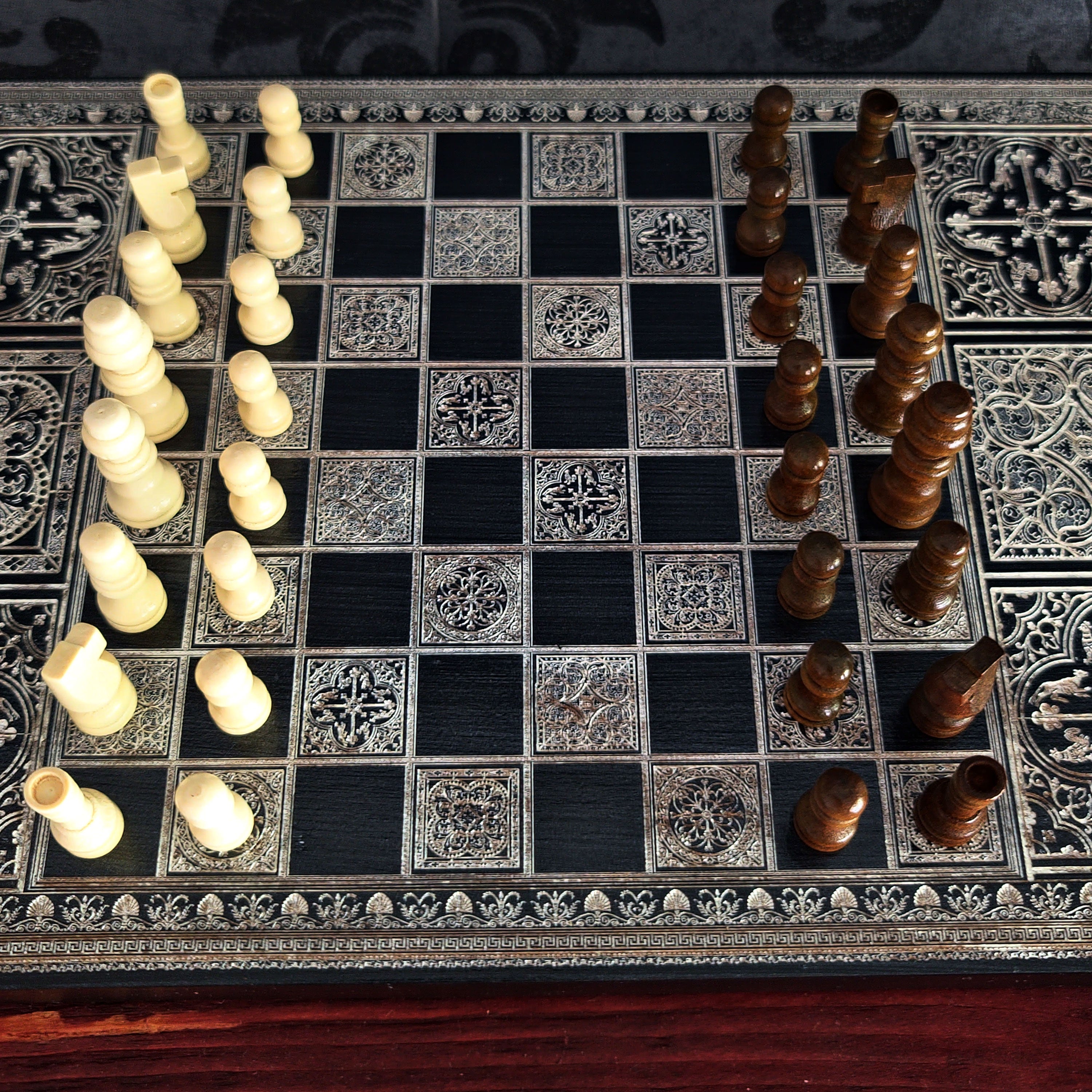 An ornate chess set with a black and silver patterned board, featuring cream-colored pawns on one side and dark brown wooden pieces on the other, photographed from a top-down angle.