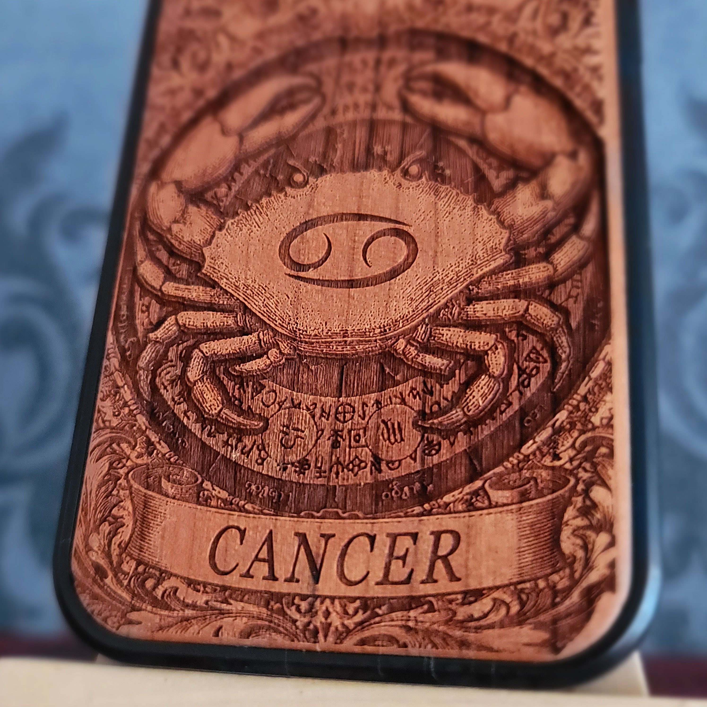 A close-up of a wooden phone case featuring an engraved Cancer zodiac symbol and the word 'CANCER' on a dark background.