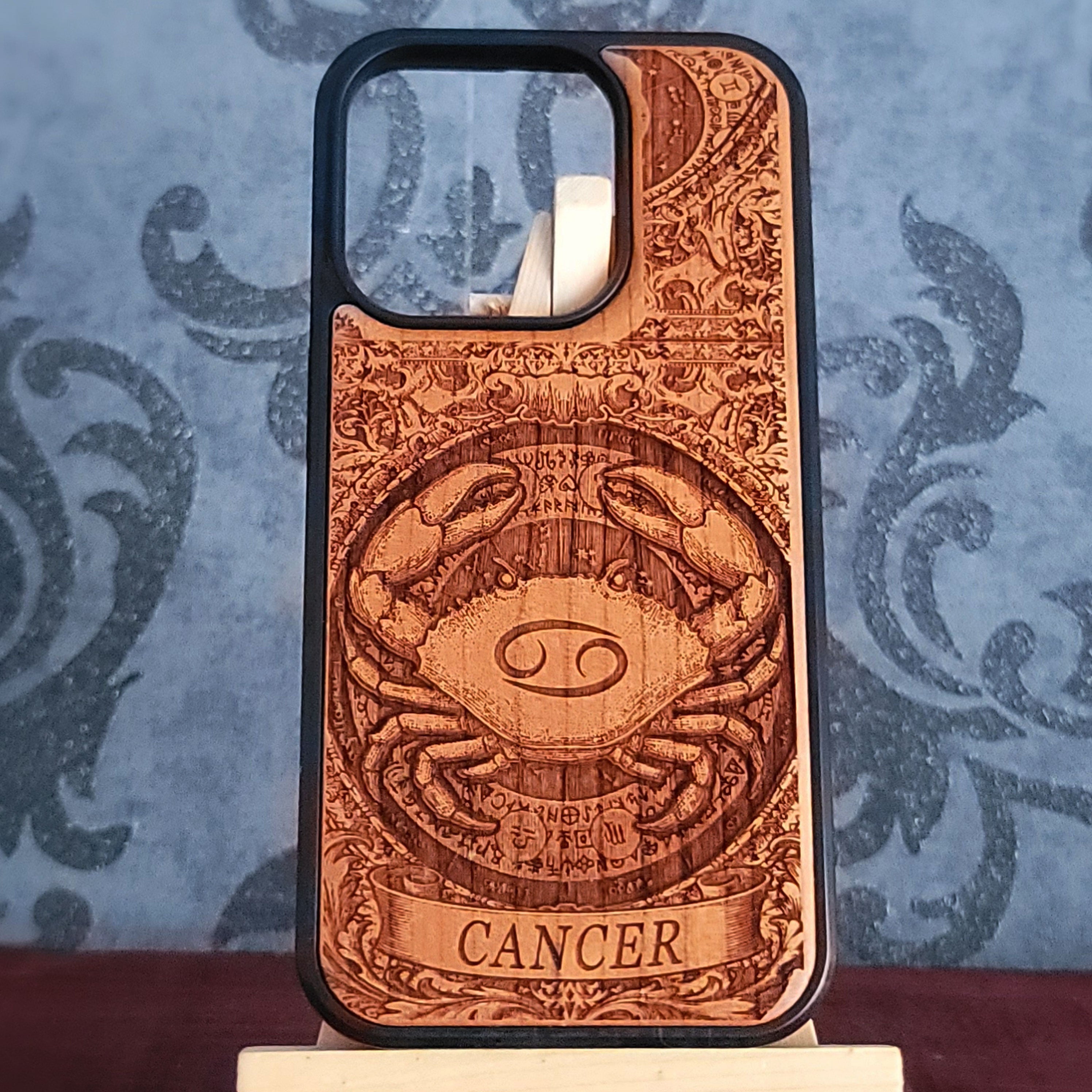 A wooden phone case with a detailed laser-engraved Cancer zodiac sign, displayed on a small stand against a blue patterned background.
