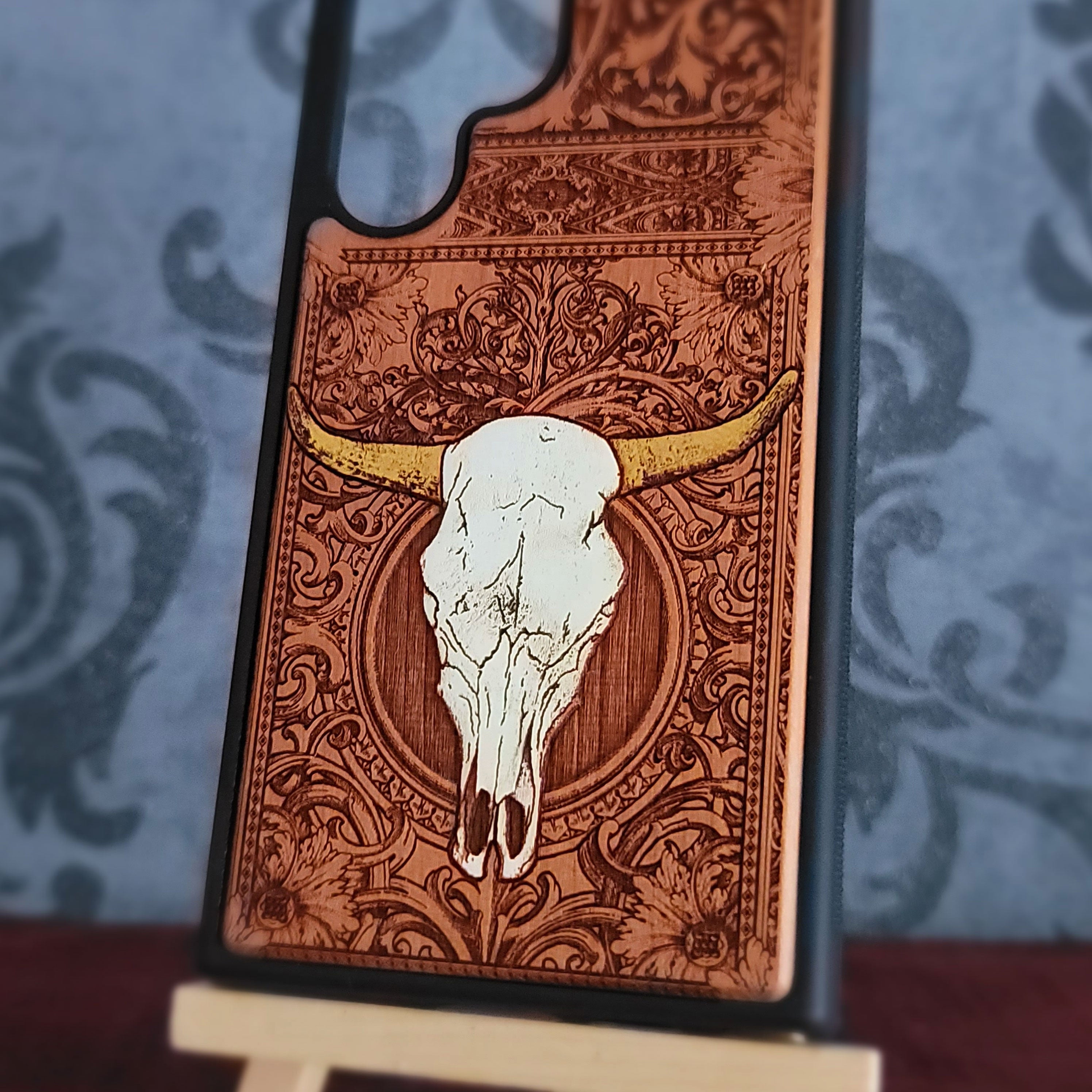 A close-up of a decorative phone case featuring a white skull with yellow horns over an ornate brown engraved pattern, shown standing on a small display stand against a blue patterned background.