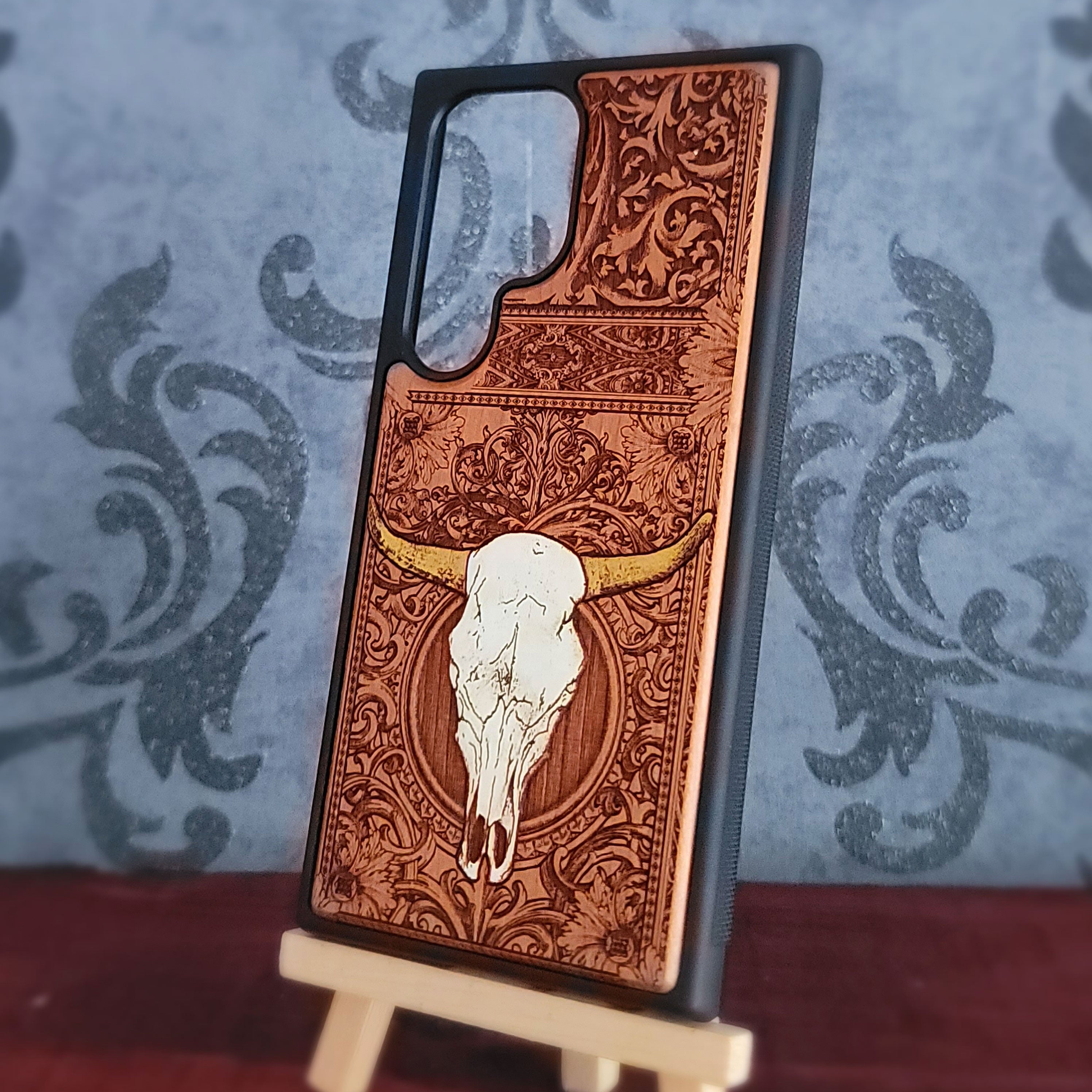 A wooden phone case with an engraved western design featuring a white skull and gold horns, displayed on a small wooden stand against a blue damask patterned background.