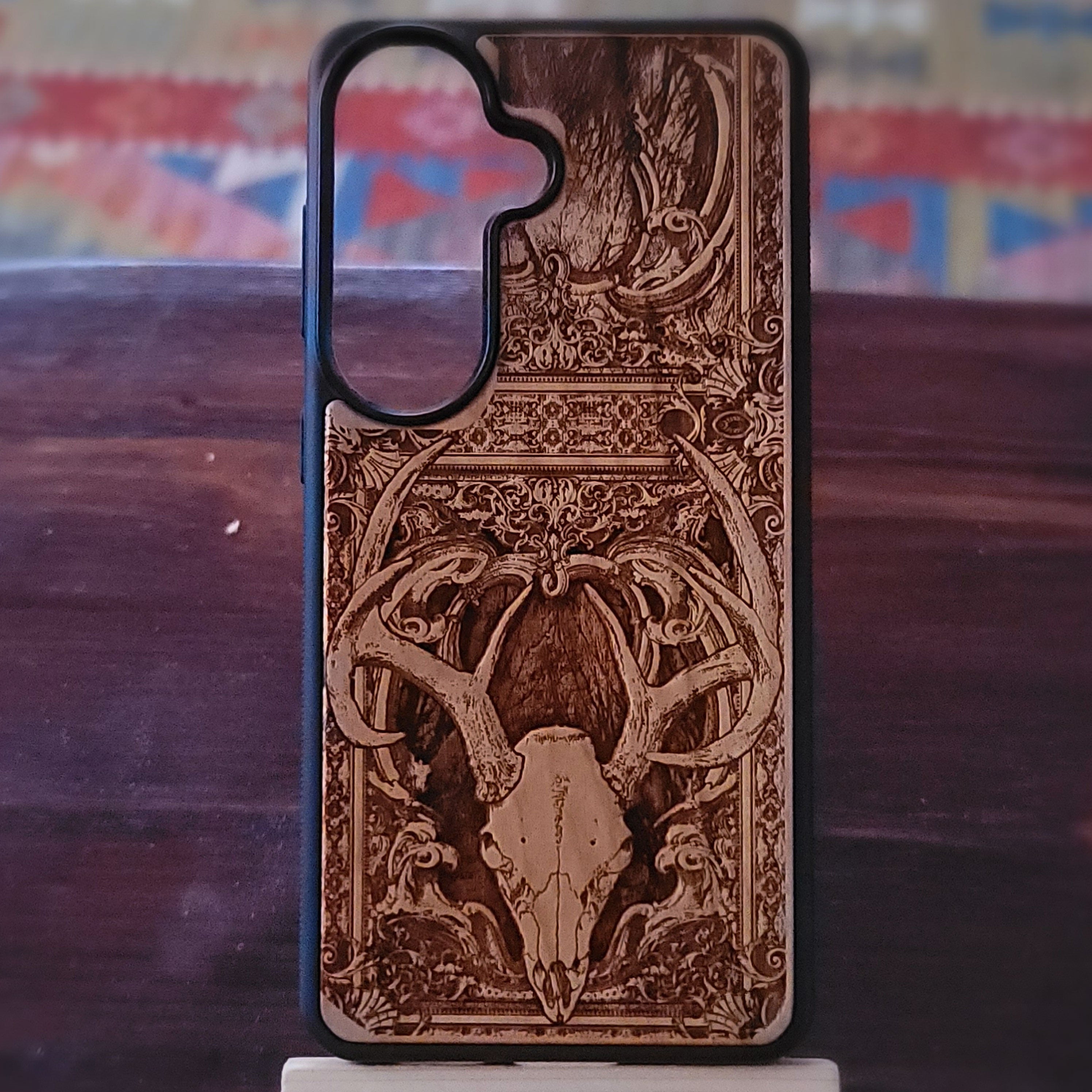 A wooden phone case with a detailed laser-burned design of a deer skull and antlers, displayed on a dark wooden surface.