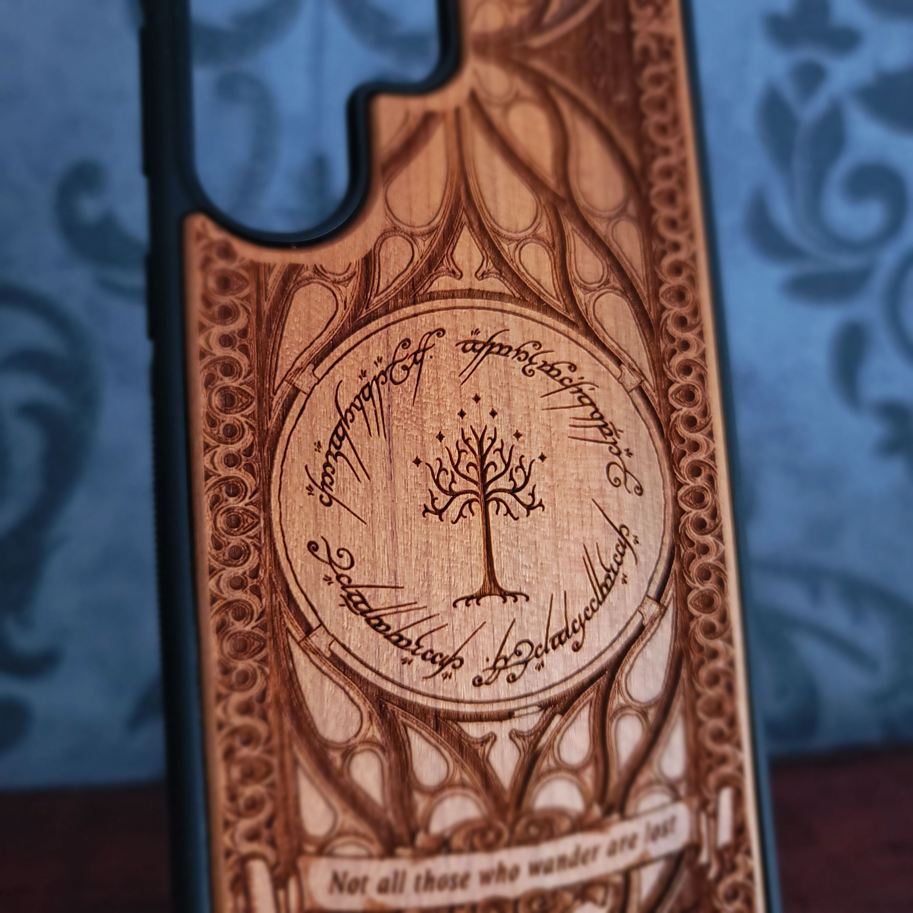 A close-up of a wooden phone case featuring an engraved circular tree design with Elvish script and the quote 'Not all those who wander are lost', shown against a blue patterned background.