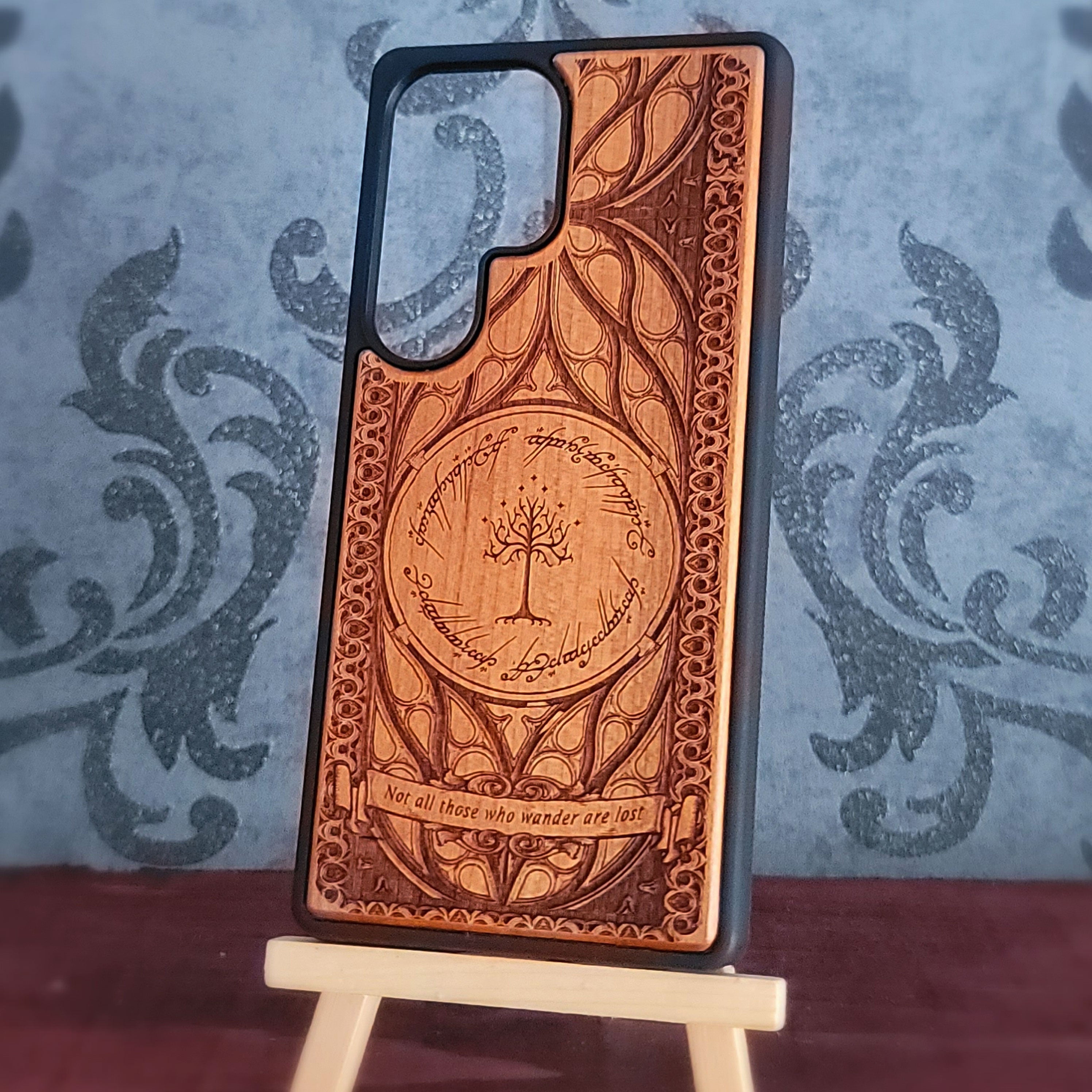 A wooden phone case with a dark gray bumper edge, featuring an engraved design inspired by The Lord of the Rings, displayed on a small wooden stand against a patterned blue-gray background.