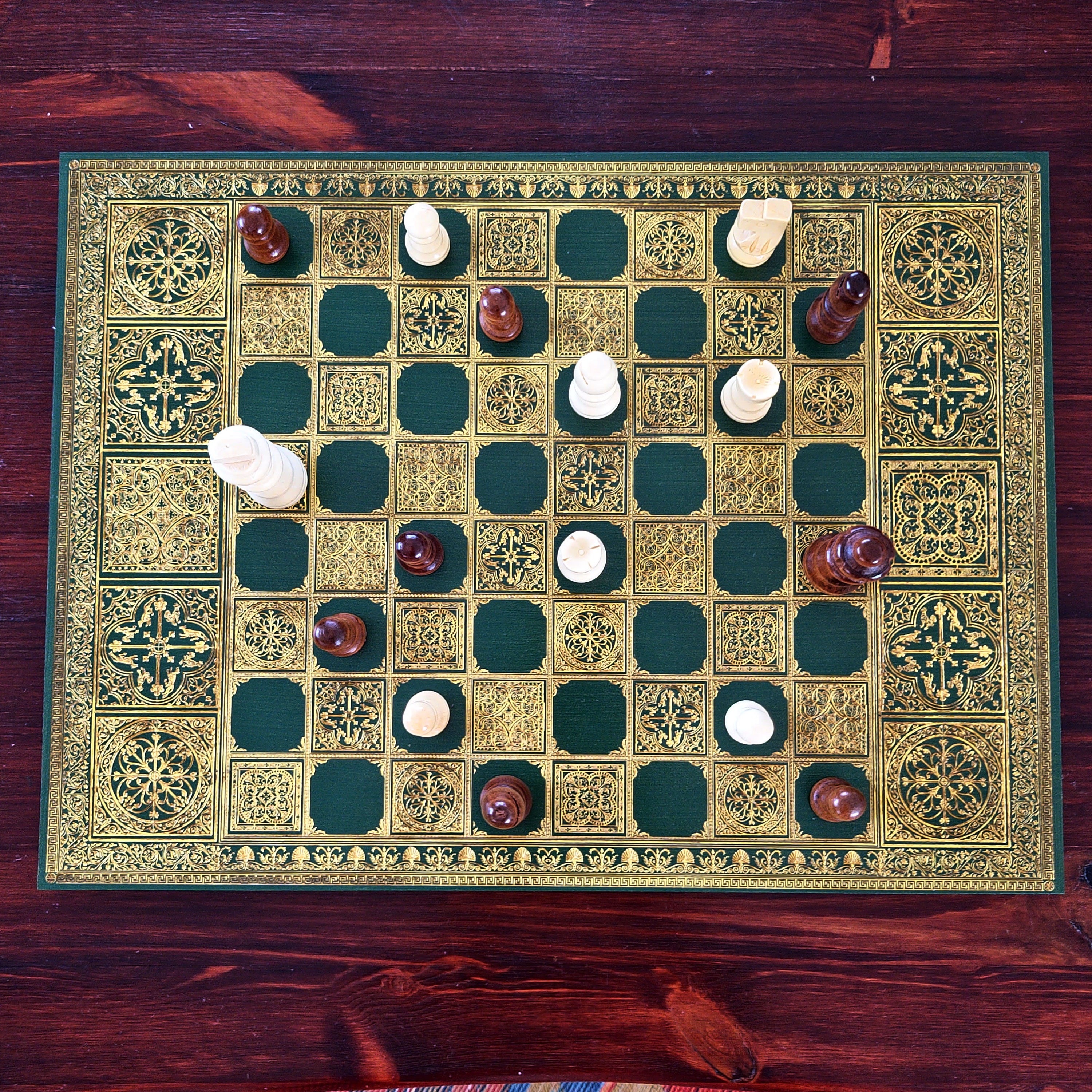 An ornate green and gold chess board set on a dark wooden table, with white and brown pieces arranged to play. The board features intricate geometric patterns and a decorative border.