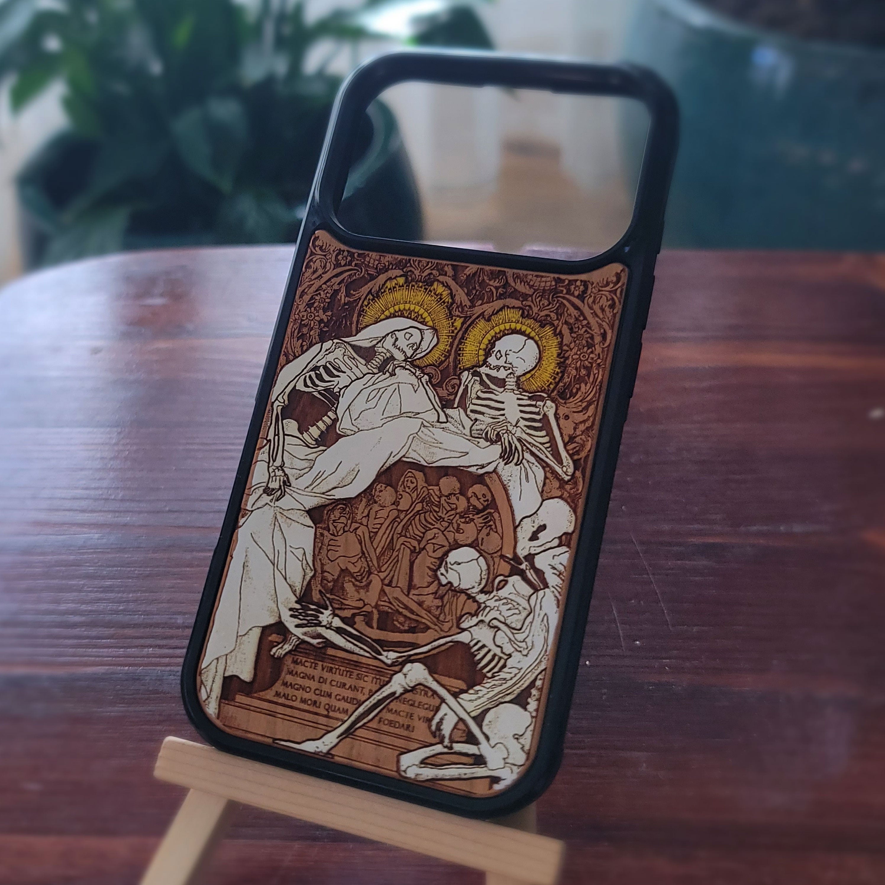 A phone case with a black bumper and a detailed medieval-style print of skeletons and religious figures, displayed on a small wooden stand on a dark wooden table.