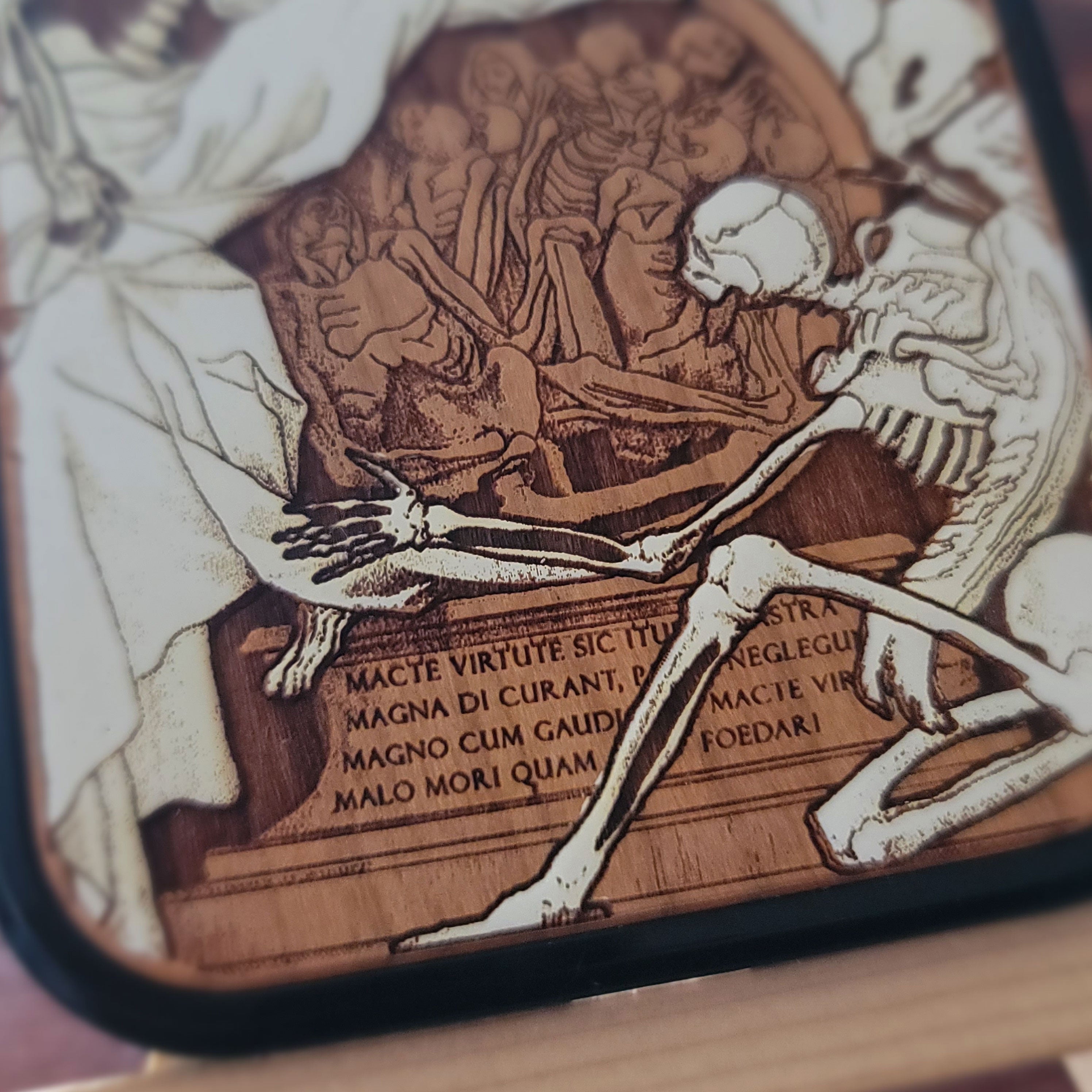 Close-up of a square wooden plaque or coaster with a dark border, featuring an engraved or burned wood illustration of a skeleton beside a figure and Latin text.