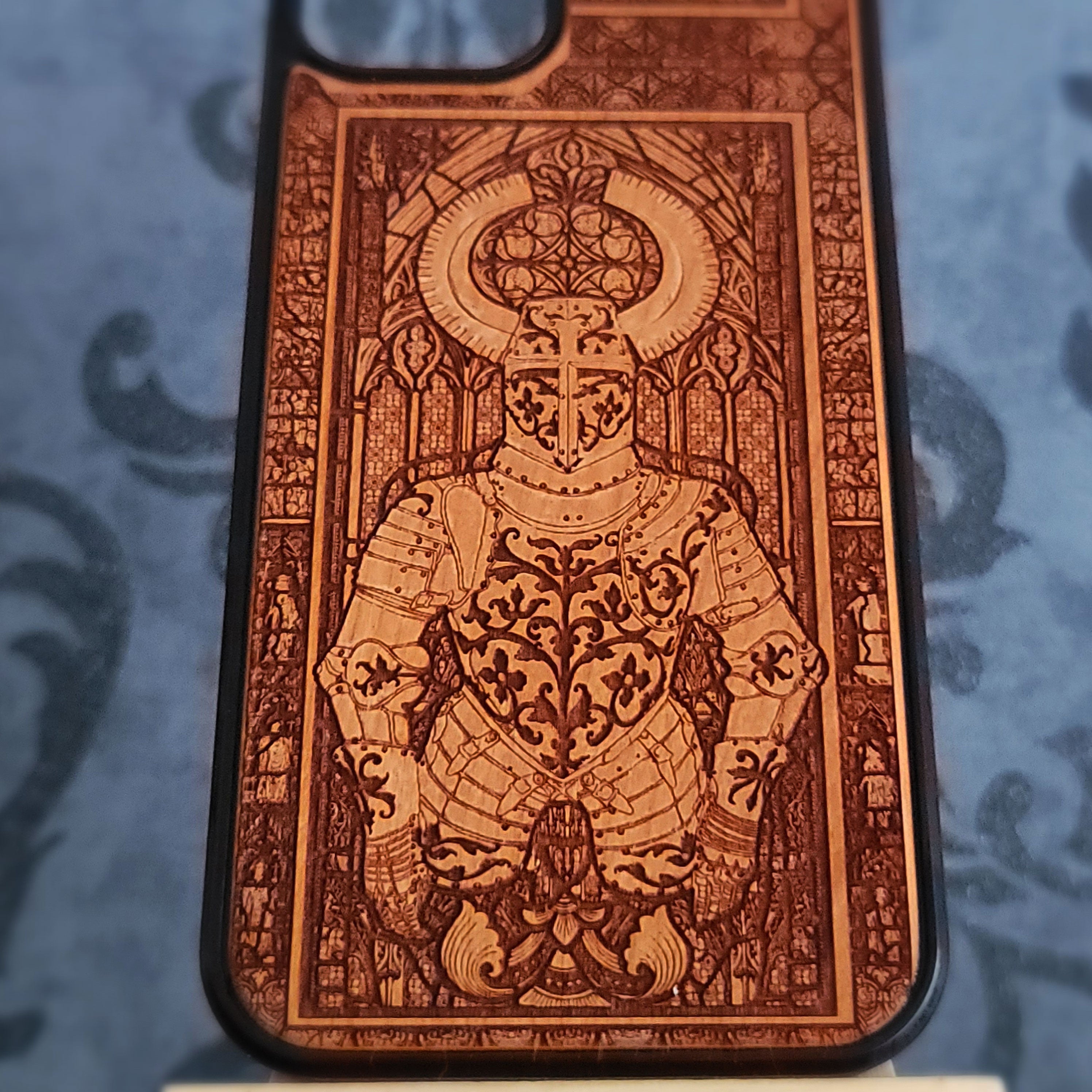 A close-up of a phone case featuring an intricate laser-engraved design of a knight in armor with a halo, set against a stained-glass-style background. The case has a warm brown wood-grain finish with a black bumper edge,