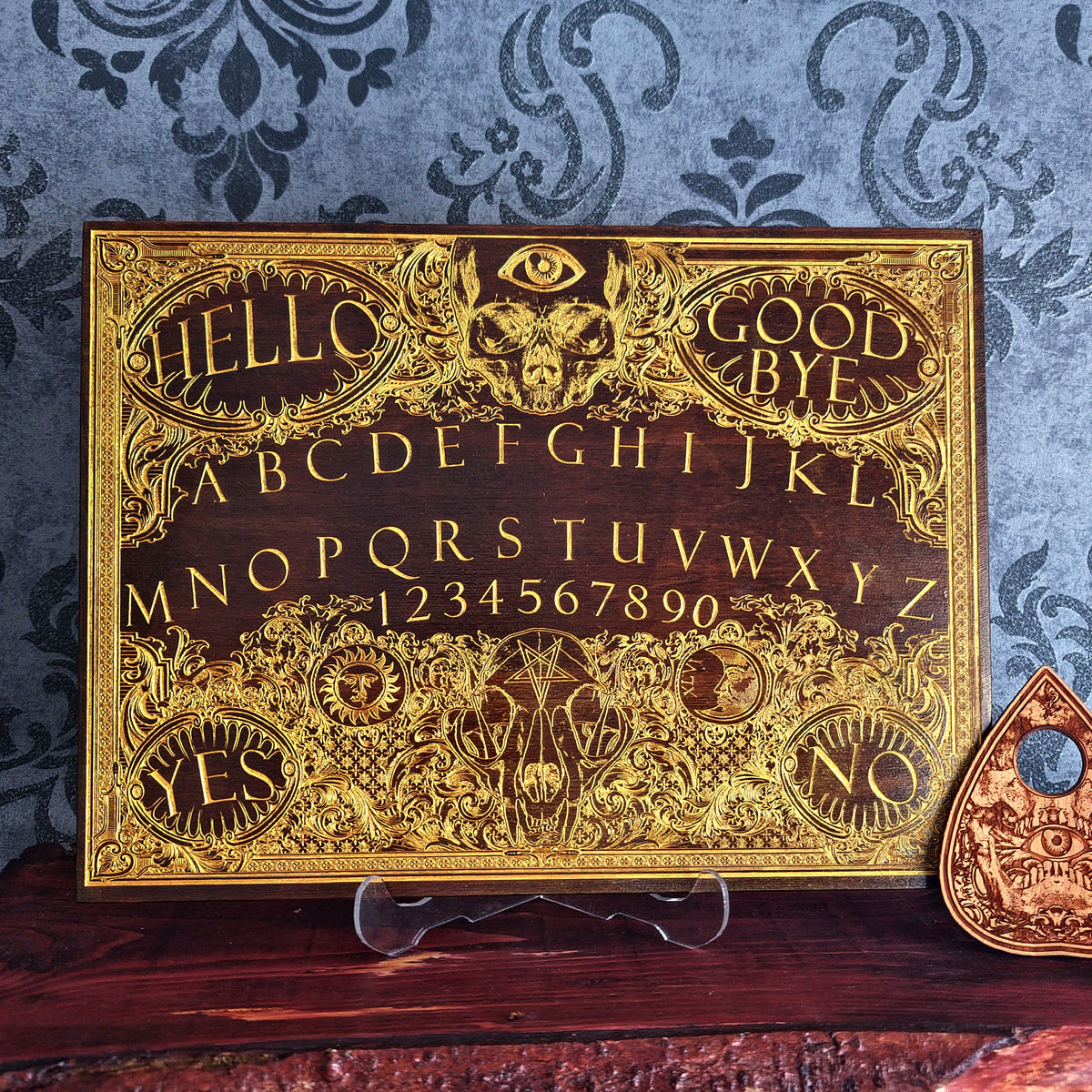 An ornate gold and dark wood Ouija board with intricate filigree details, A matching planchette rests to the right.