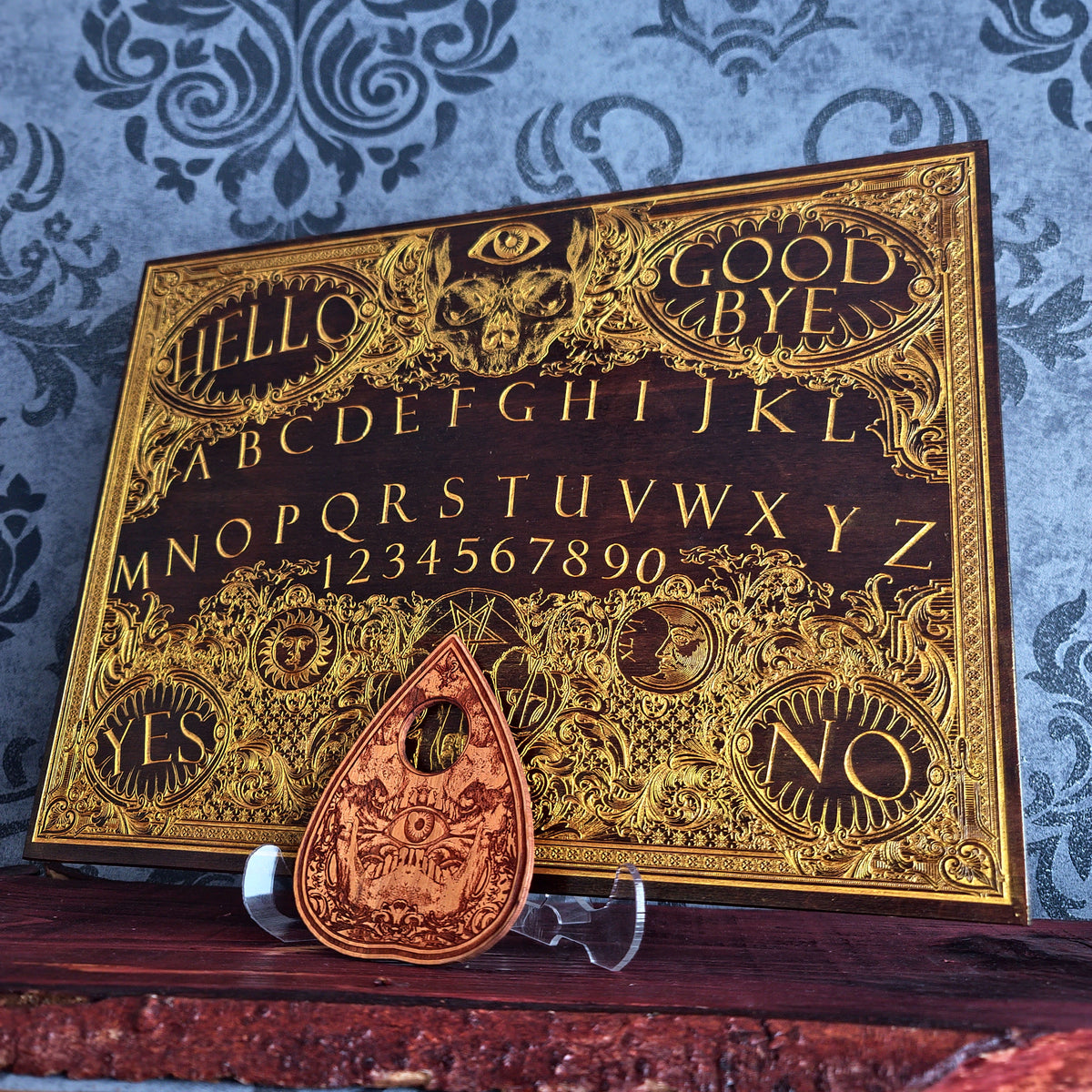 A dark wood Ouija board with ornate gold foil lettering and symbols, displayed on a clear stand with a matching teardrop-shaped copper planchette in front