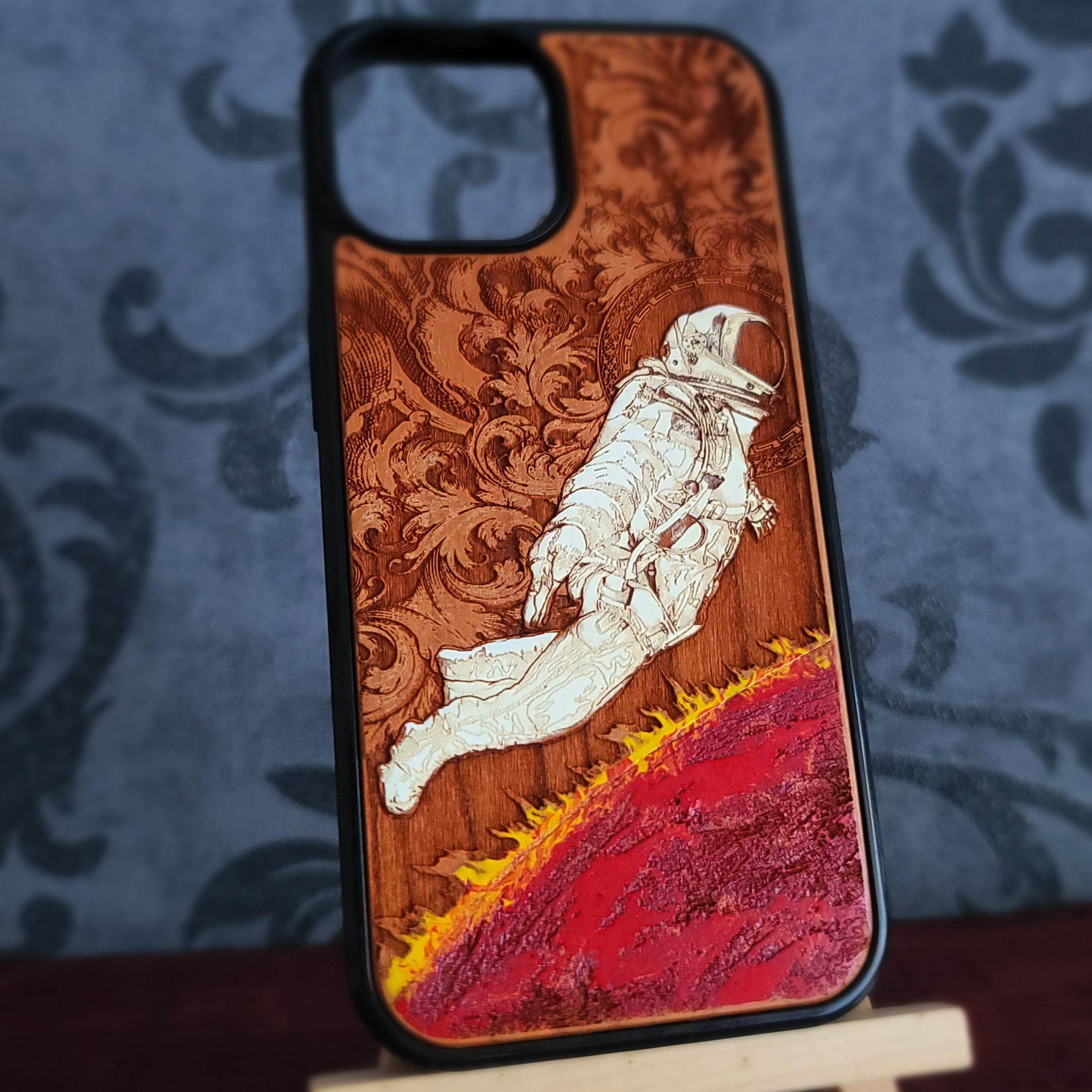 A wooden phone case with a laser-engraved design of an astronaut floating above a red and yellow planetary landscape, set against a dark damask patterned background.