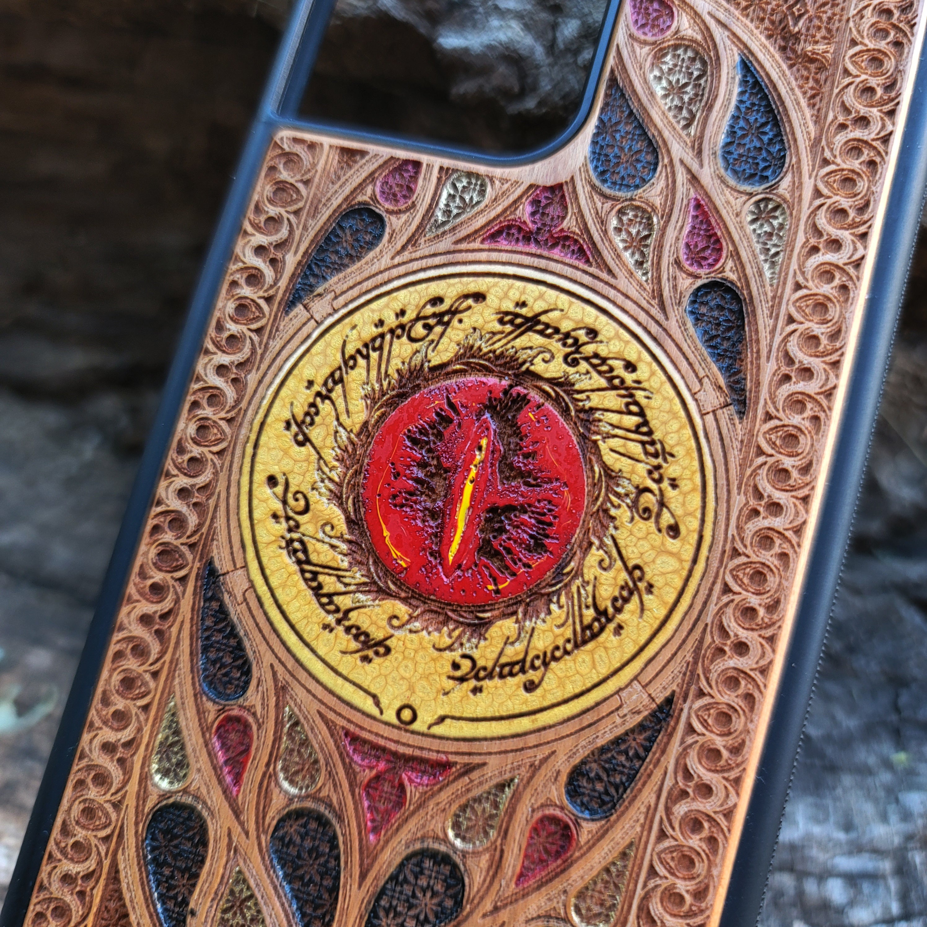 "Hand-painted wood phone case featuring the Eye of Sauron surrounded by intricate Elvish script and vibrant colored patterns. This laser-engraved and detailed design offers a unique blend of artistry and durability. Compatible with iPhone , 15, 14, 13, 12, 11, Pro Max, Mini, and Samsung Galaxy S, S24, S23, S22, S21, S20 Ultra, Plus, Note 20. Perfect for Lord of the Rings fans seeking a bold and protective accessory