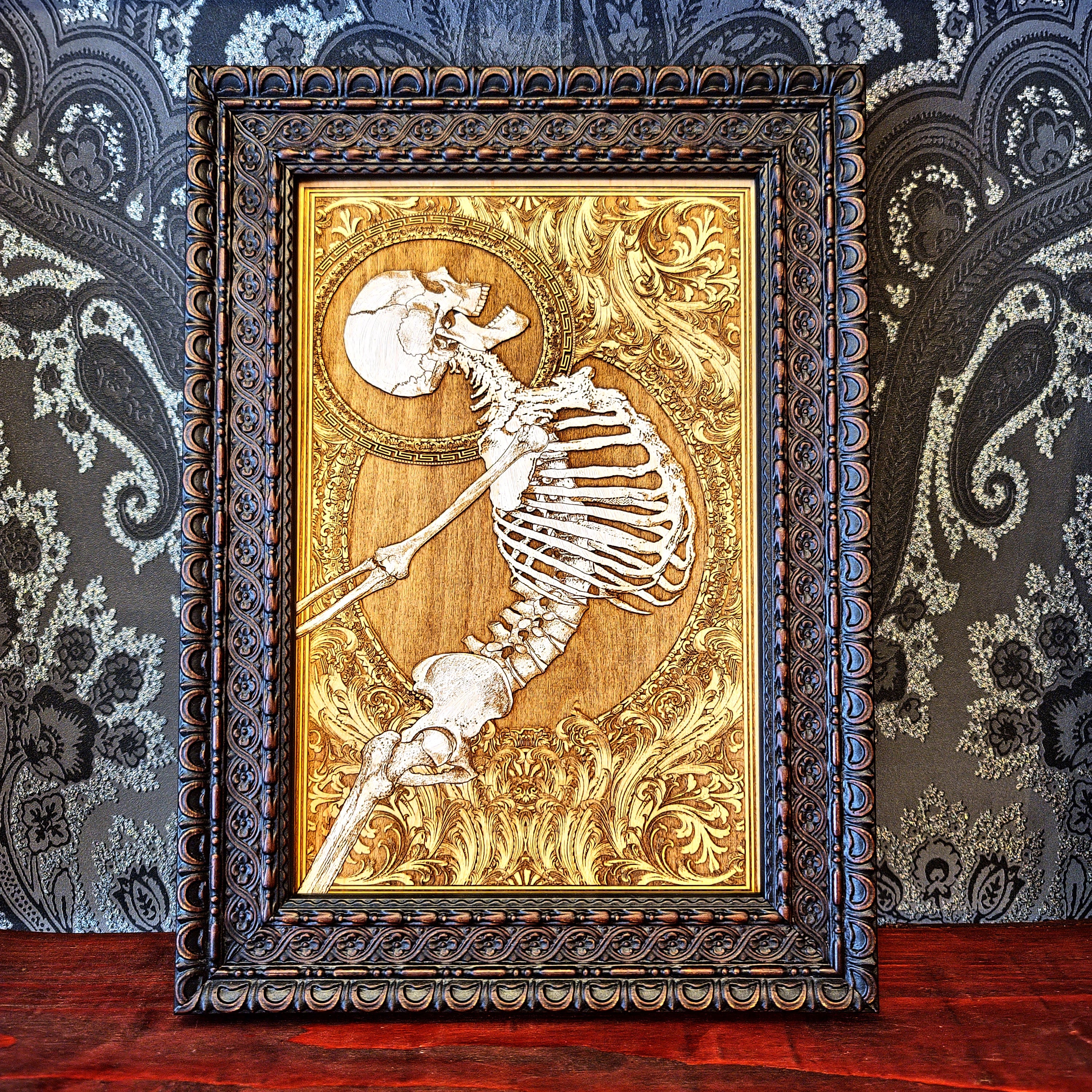 Decorative wall art with a skeleton design in a ornate frame against a patterned wall.