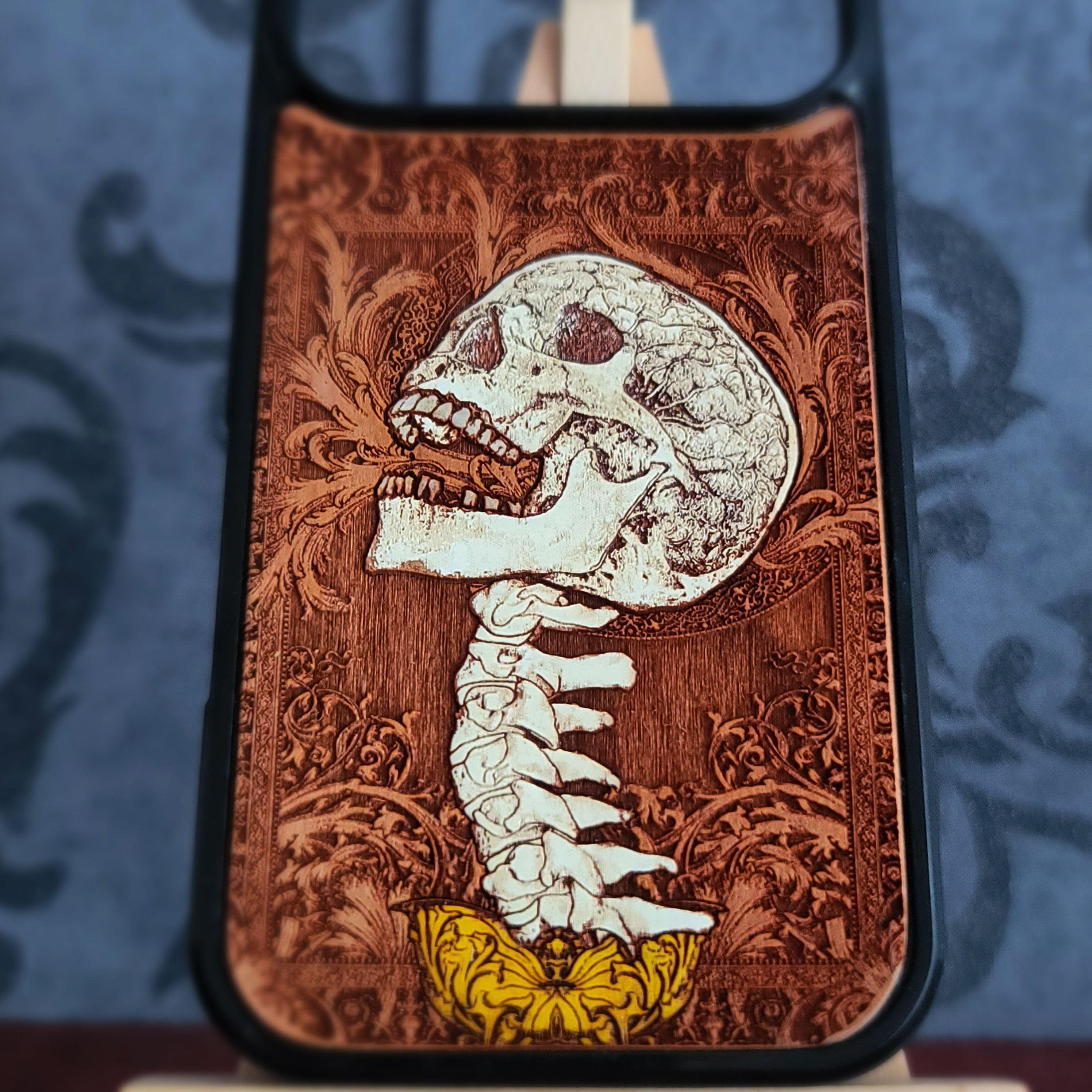 A close-up of a phone case featuring a detailed laser-engraved design of a skull and spine in white and brown, with a yellow butterfly accent at the bottom, set against a dark blue patterned background.