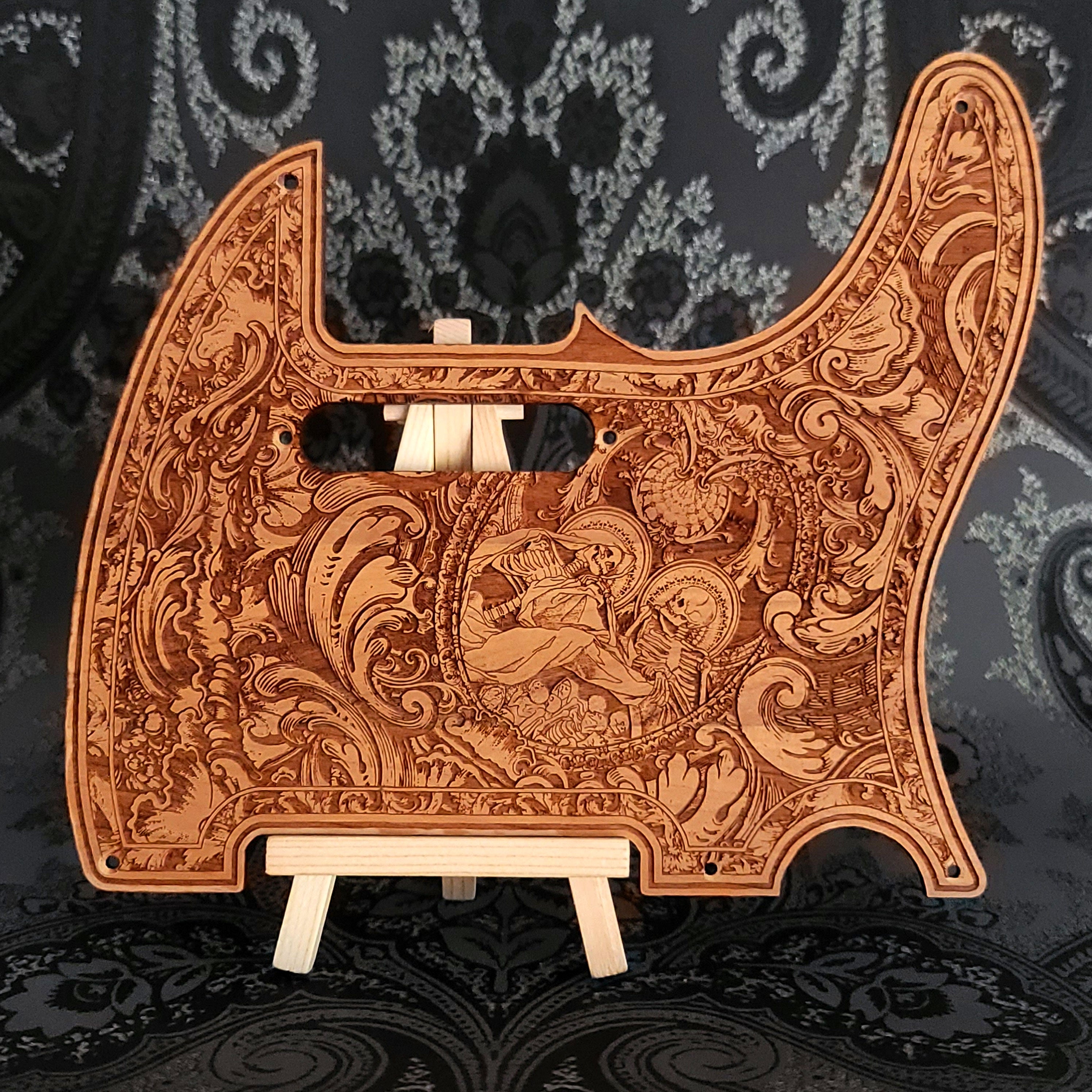 A wooden guitar pickguard with an intricate laser-etched design featuring a skeletal figure and floral patterns.