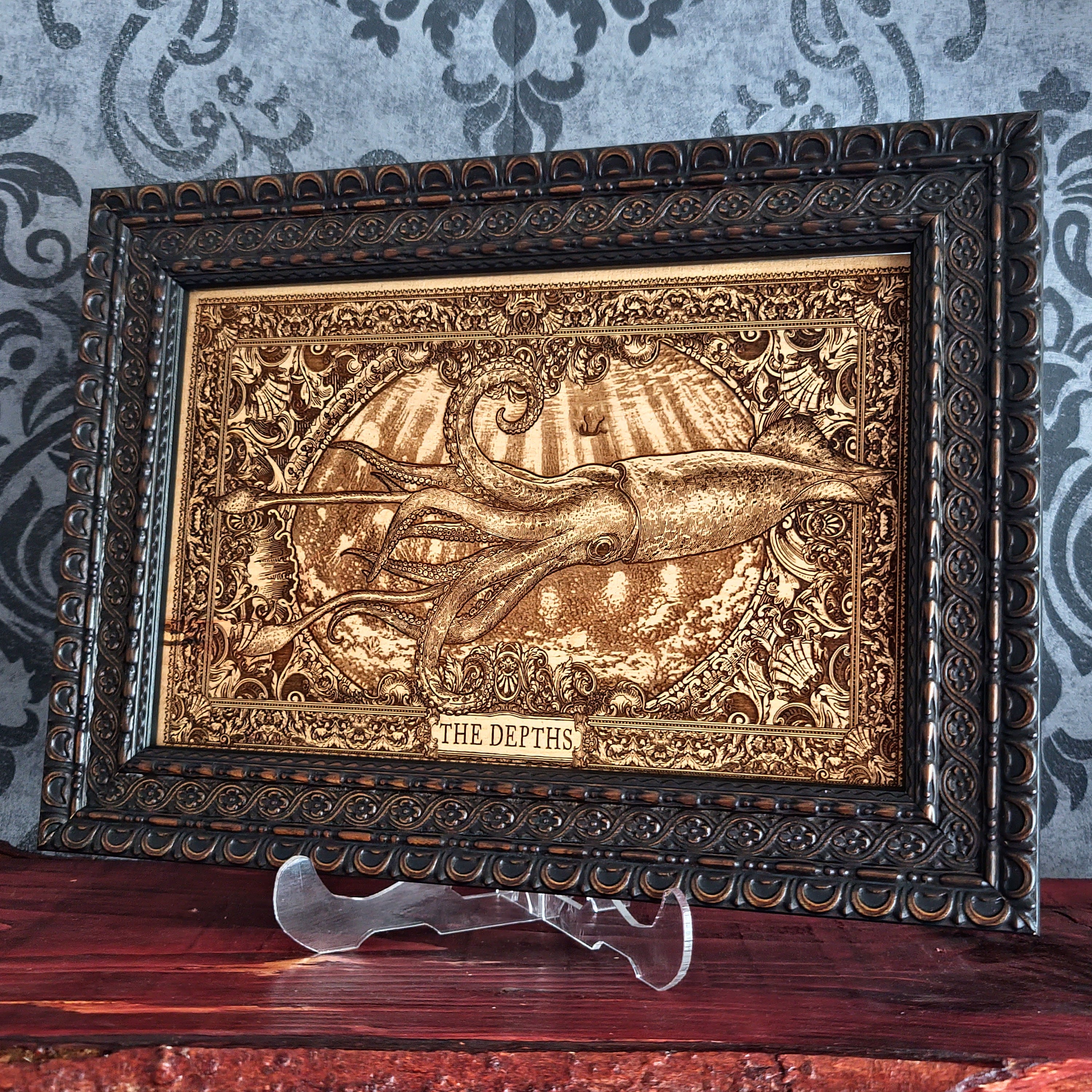 An ornate framed decorative art piece featuring a raised octopus emerging from water, with the words 'THE DEPTHS' at the bottom. The piece has a dark carved frame and is displayed on a clear stand against a patterned wallpaper background.