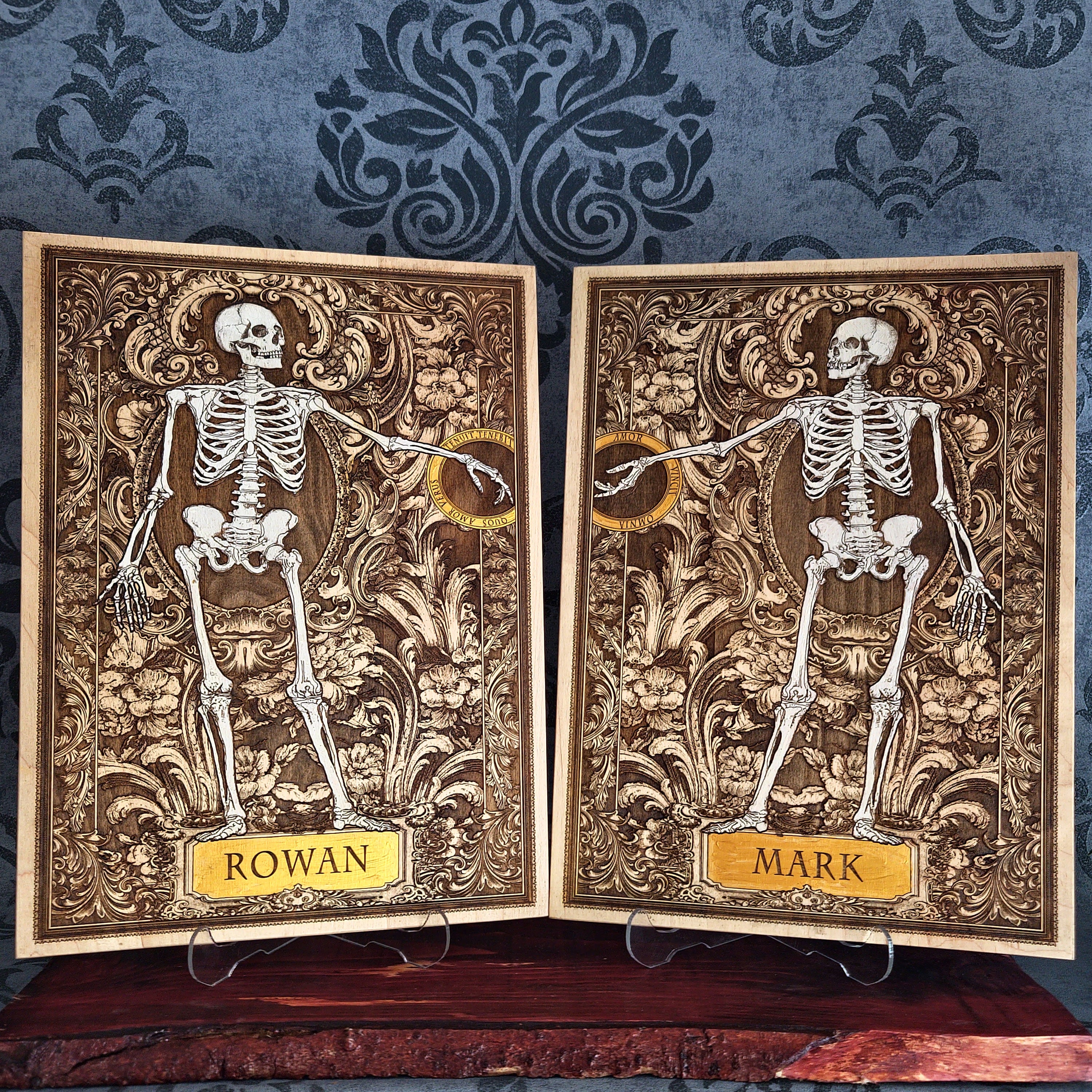 Two personalized gothic-style skeleton cards displayed side-by-side on small clear stands. The cards feature detailed ornate brown and gold backgrounds with white skeleton illustrations, one labeled 'ROWAN' and the other 'MARK'.