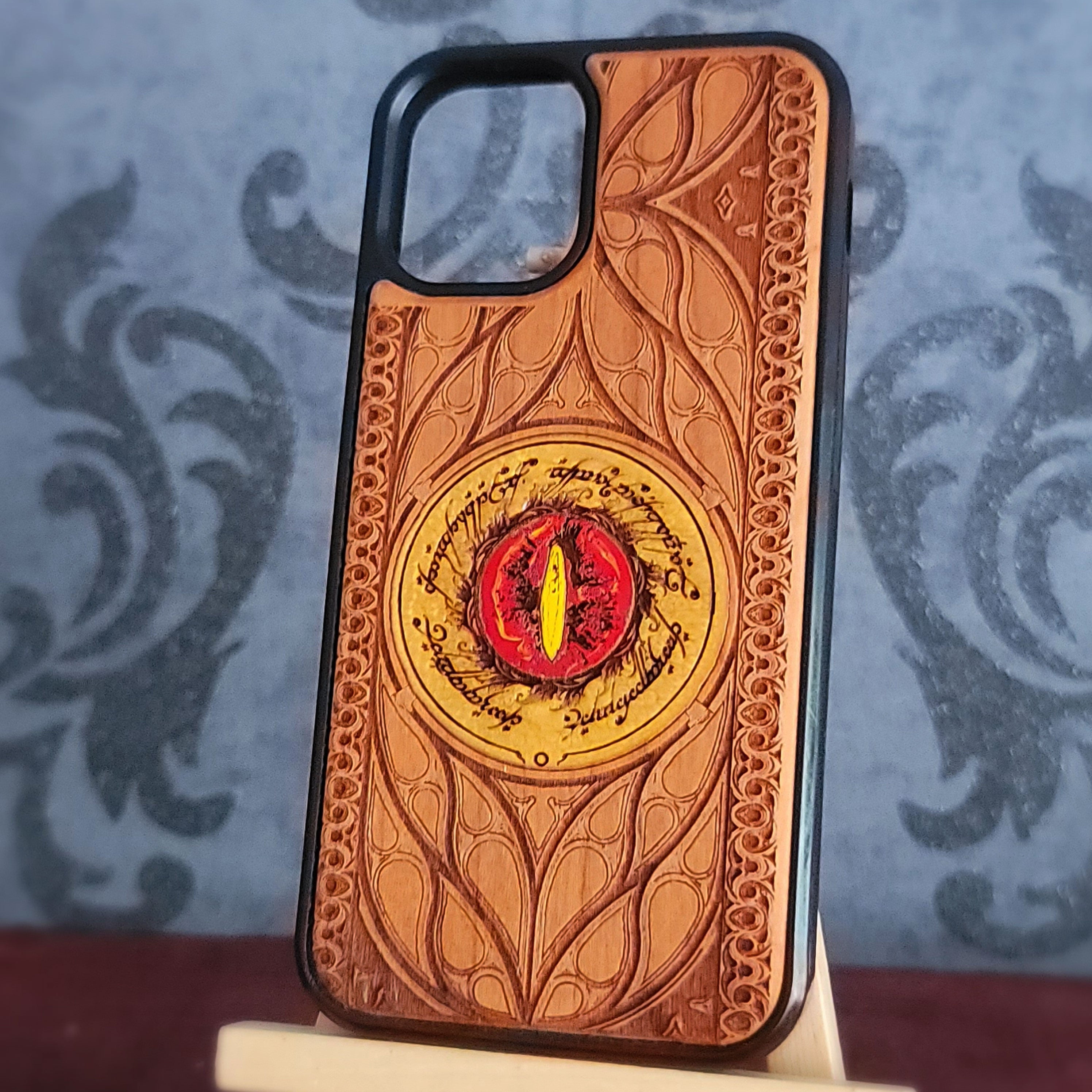 A phone case featuring an embossed brown leaf pattern and a central Elvish ring design with a red eye and yellow iris, displayed on a small stand against a patterned gray background.