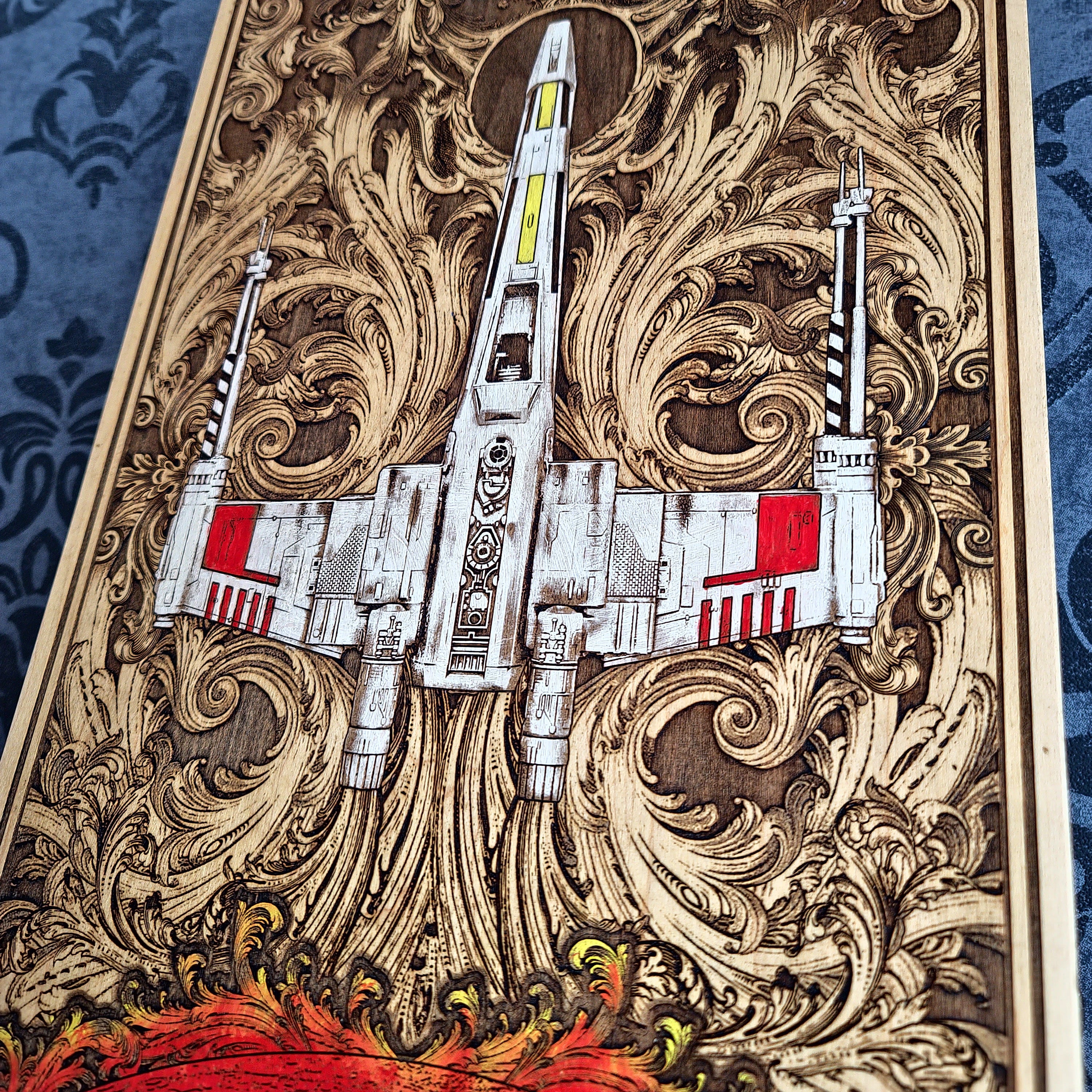 A close-up of a decorative wooden panel or artwork featuring a white X-wing style spacecraft over ornate swirling wood-carved patterns, with red accents and a blue patterned fabric visible at the sides.