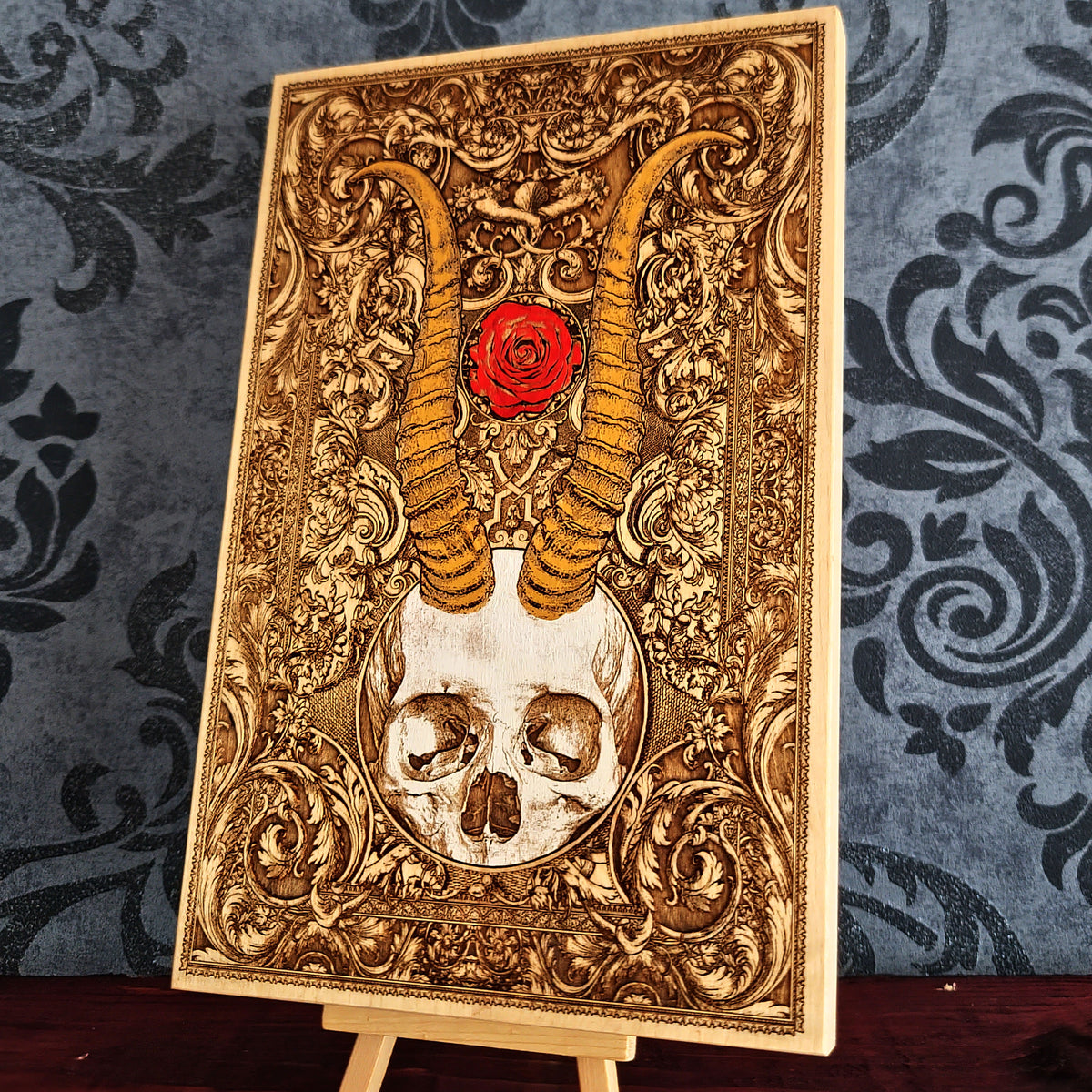 An ornate rectangular decorative panel or plaque on a small easel stand, featuring a white skull at the bottom, two curved golden horns, a red rose near the top, and intricate gold scrollwork against a dark patterned backdrop.