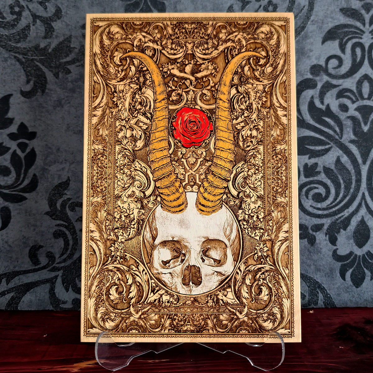 A rectangular decorative plaque or art print with an ornate gold-toned baroque-style design, featuring a skull at the bottom, large curved horns rising upward, and a red rose near the top center. 