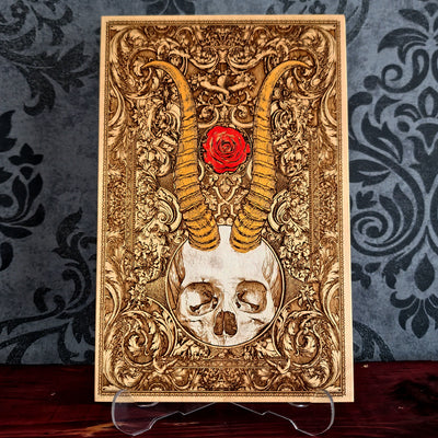 A rectangular decorative plaque or art print with an ornate gold-toned baroque-style design, featuring a skull at the bottom, large curved horns rising upward, and a red rose near the top center. 