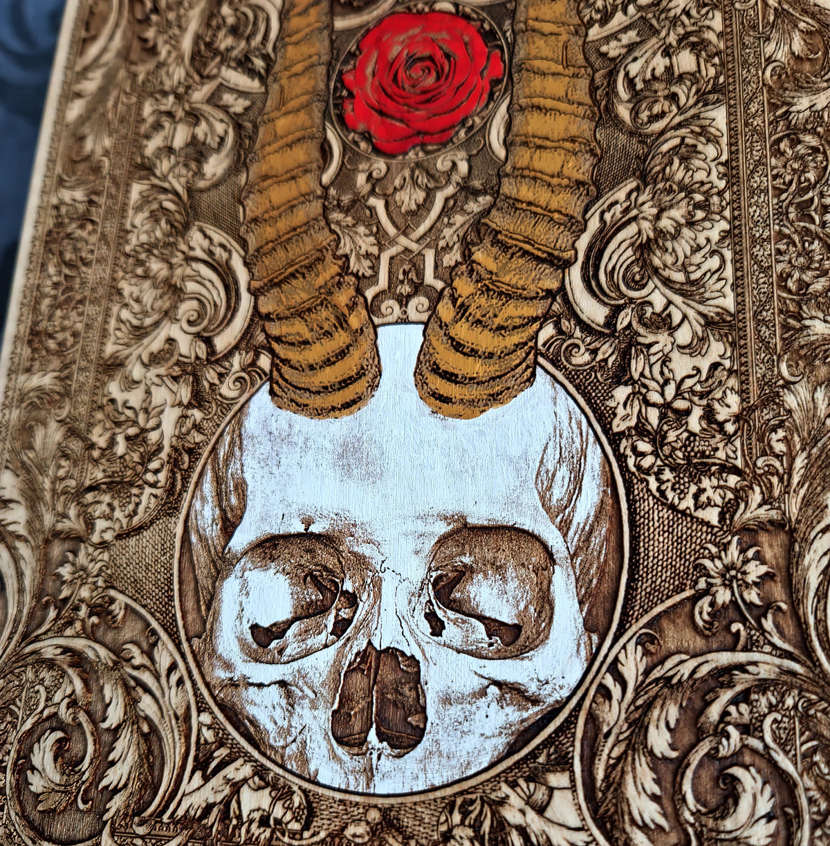 Close-up of an ornate decorative artwork featuring a white skull in an oval frame, tan ram horns, a red rose, and intricate carved floral scrollwork in brown and beige tones.