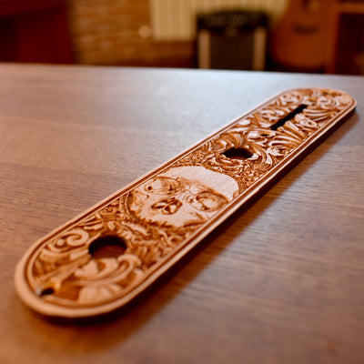 Laser-engraved dark wood control plate for a Telecaster guitar, featuring a stylised skull motif with intricate swirling background, warm brown-black tones and mounting holes visible at bottom edge.