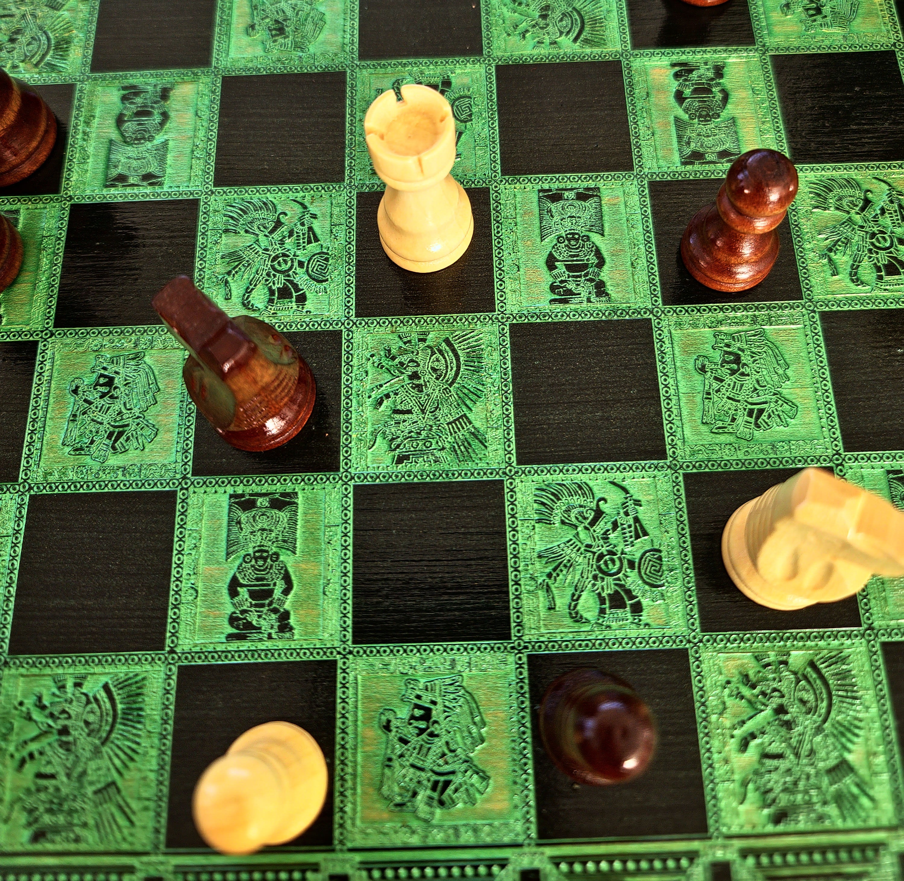 A close-up of a decorative chessboard with green and black squares, each square featuring an engraved Aztec-style warrior design. Several wooden chess pieces are scattered across the board, including a light cream rook, a dark reddish-brown pawn, and other pieces in similar finishes.