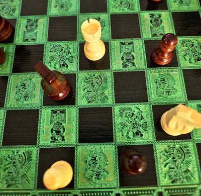 A close-up of a decorative chessboard with green and black squares, each square featuring an engraved Aztec-style warrior design. Several wooden chess pieces are scattered across the board, including a light cream rook, a dark reddish-brown pawn, and other pieces in similar finishes.