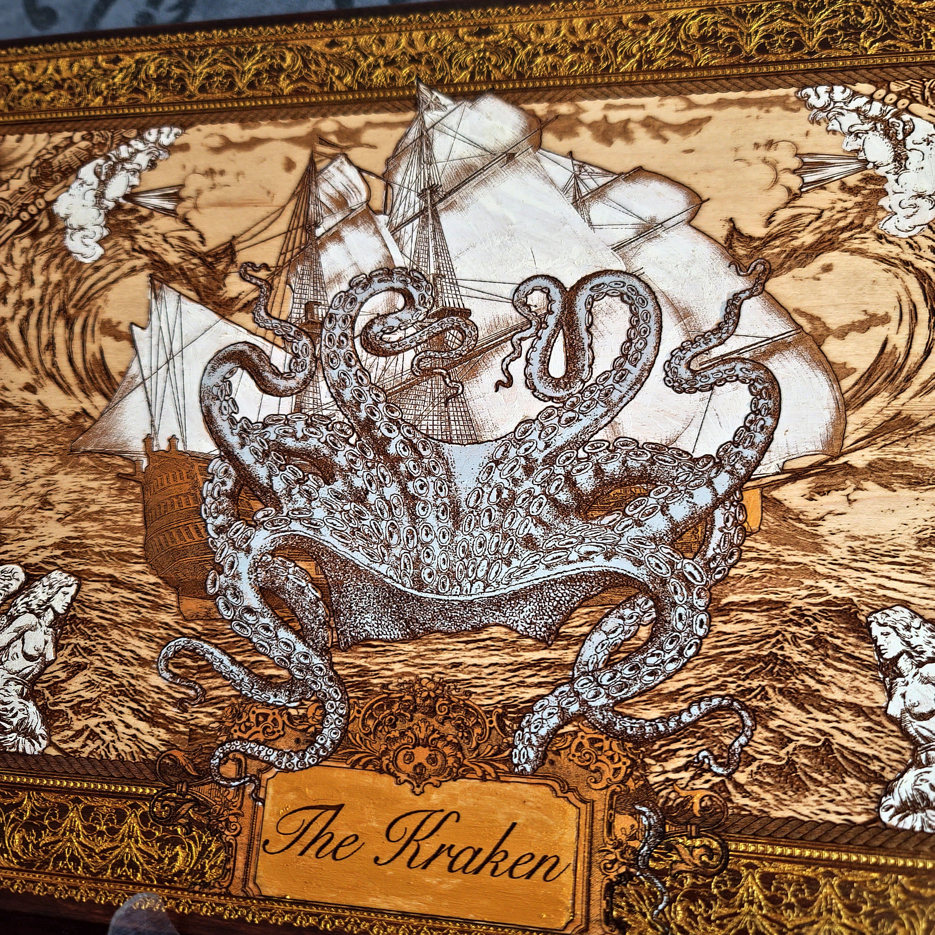 A close-up of an ornate decorative box lid featuring a detailed illustration of 'The Kraken' with a large silver-gray octopus wrapping around a white sailing ship on a brown wood-grain background, framed by an intricate gold-toned border.