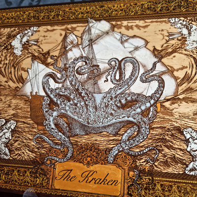 A close-up of an ornate decorative box lid featuring a detailed illustration of 'The Kraken' with a large silver-gray octopus wrapping around a white sailing ship on a brown wood-grain background, framed by an intricate gold-toned border.