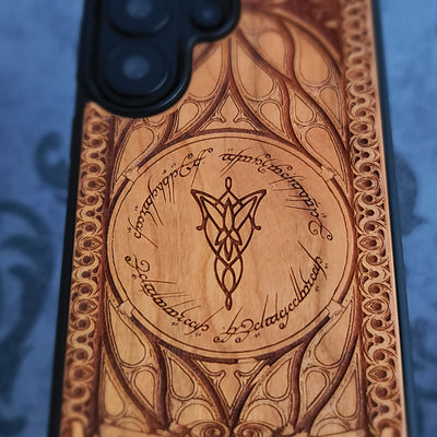 A close-up of a phone case featuring an intricate laser-engraved design inspired by The Lord of the Rings, with a central Elvish emblem and Elvish script on a light wood-grain background.