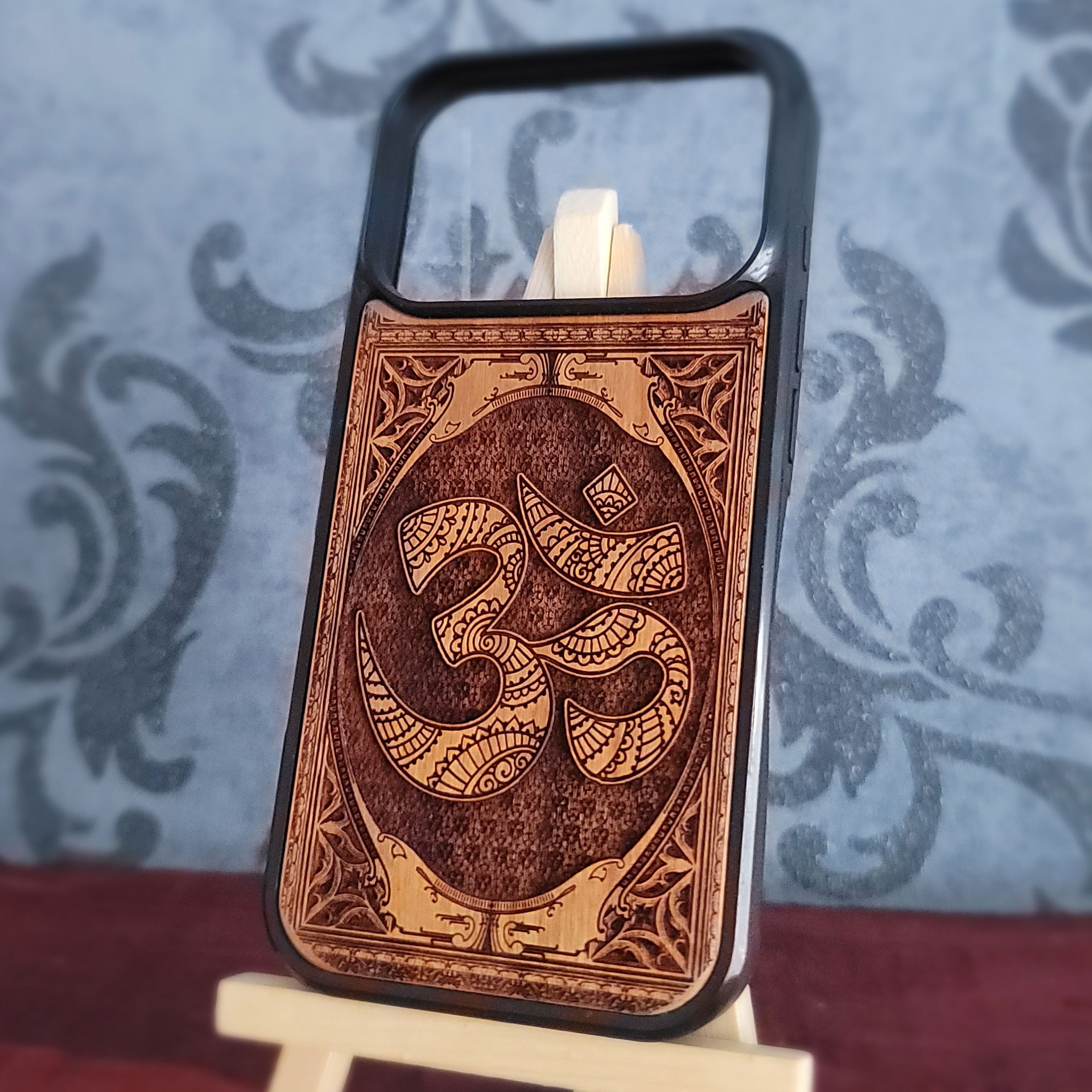 A wooden phone case with a black bumper frame, featuring a laser-engraved Om symbol and decorative border. 