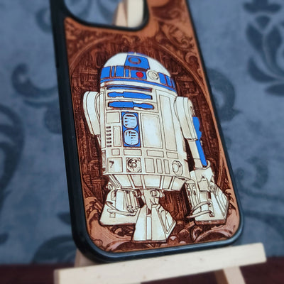 A close-up of a phone case featuring an engraved or laser-burned design of R2-D2 on a brown background, resting on a small wooden stand against a blue patterned backdrop.